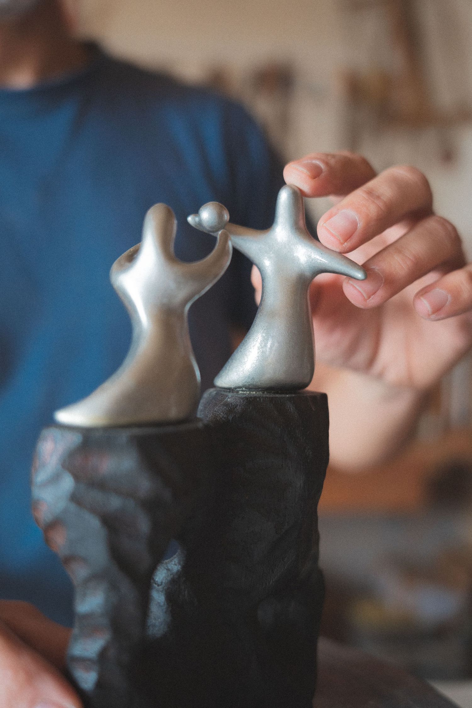 Sculptor holding small silver figurines on a dark ceramic base, depicting two abstract dancing human forms.