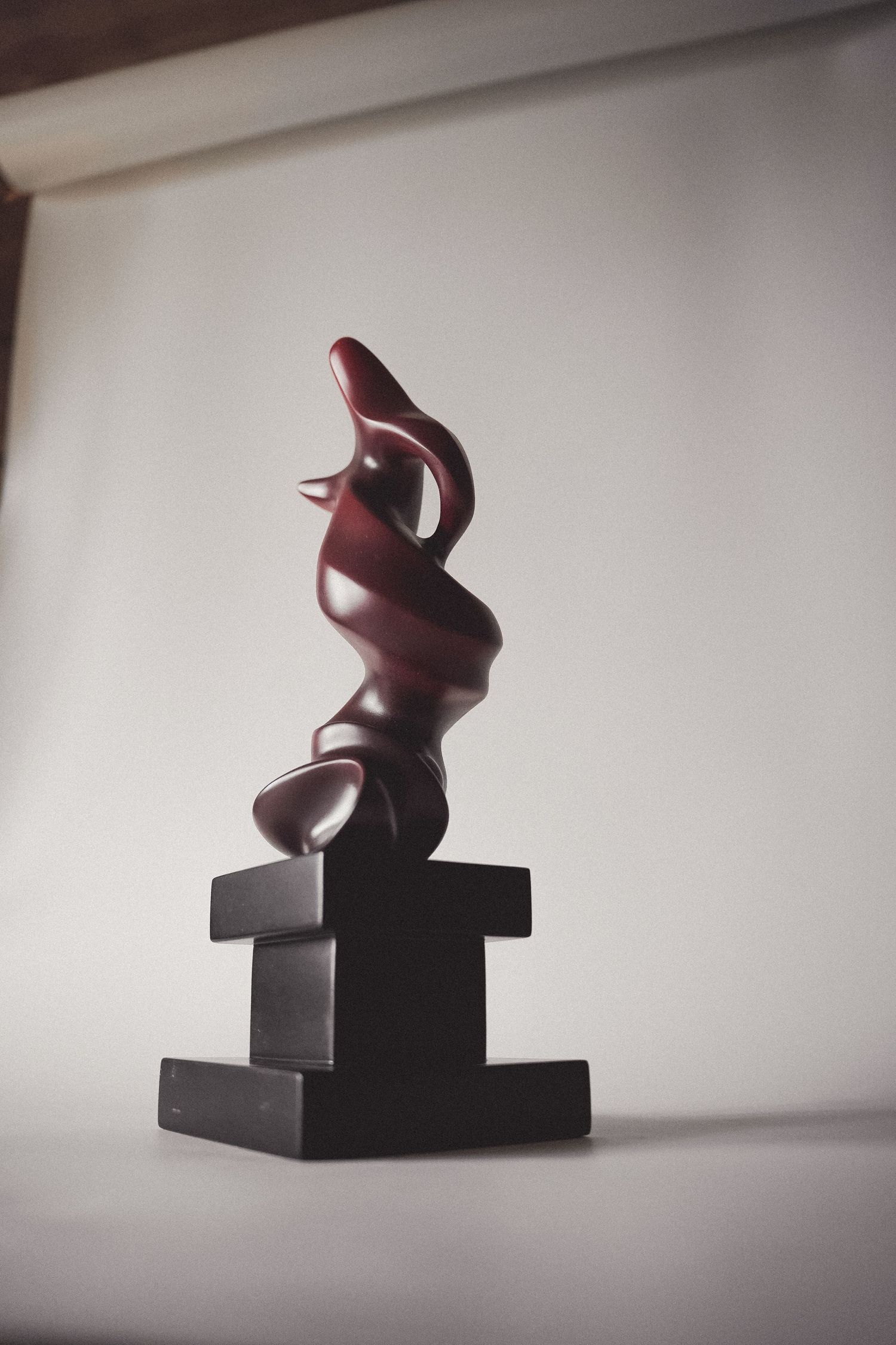 Tall dark red abstract sculpture with flowing form on a black base near a window.