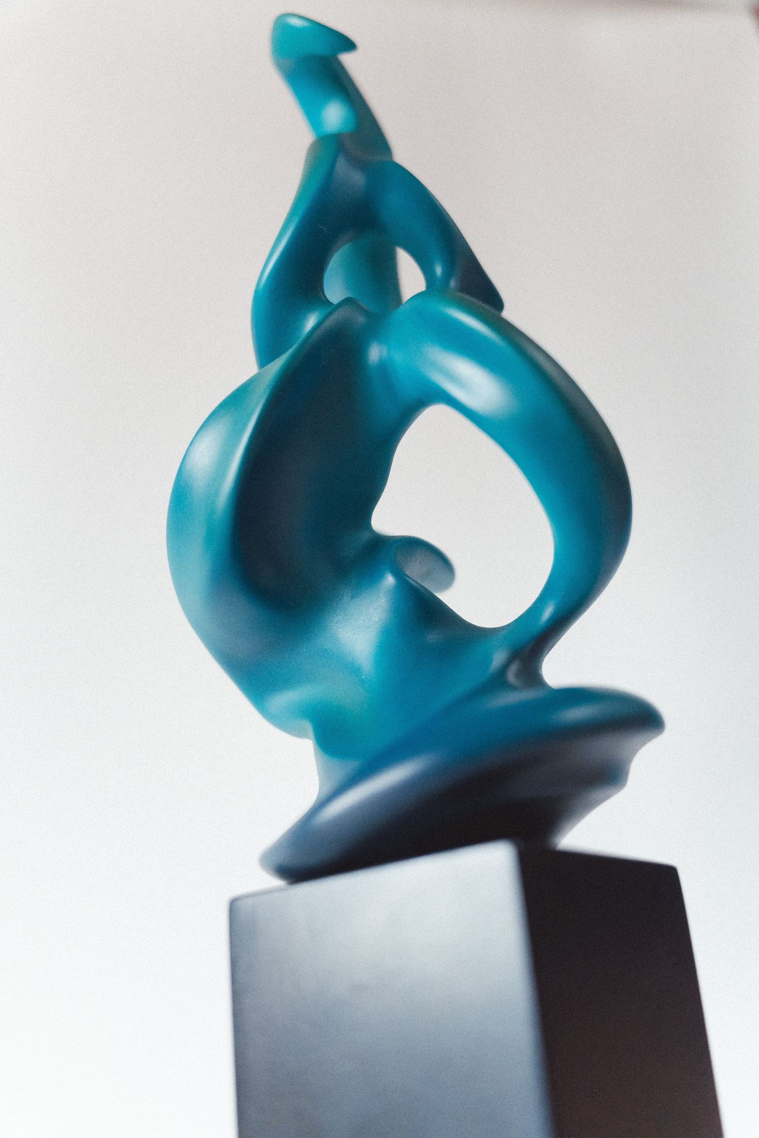 A teal abstract sculpture with fluid curves mounted on a dark square base against a white background.