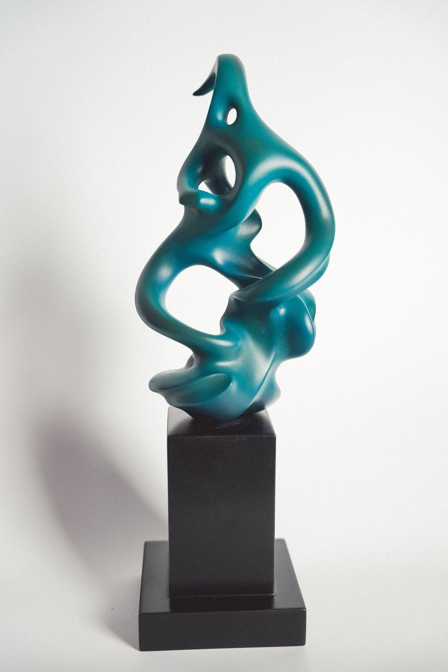 Teal abstract sculpture with fluid organic curves mounted on a black pedestal against a white background.