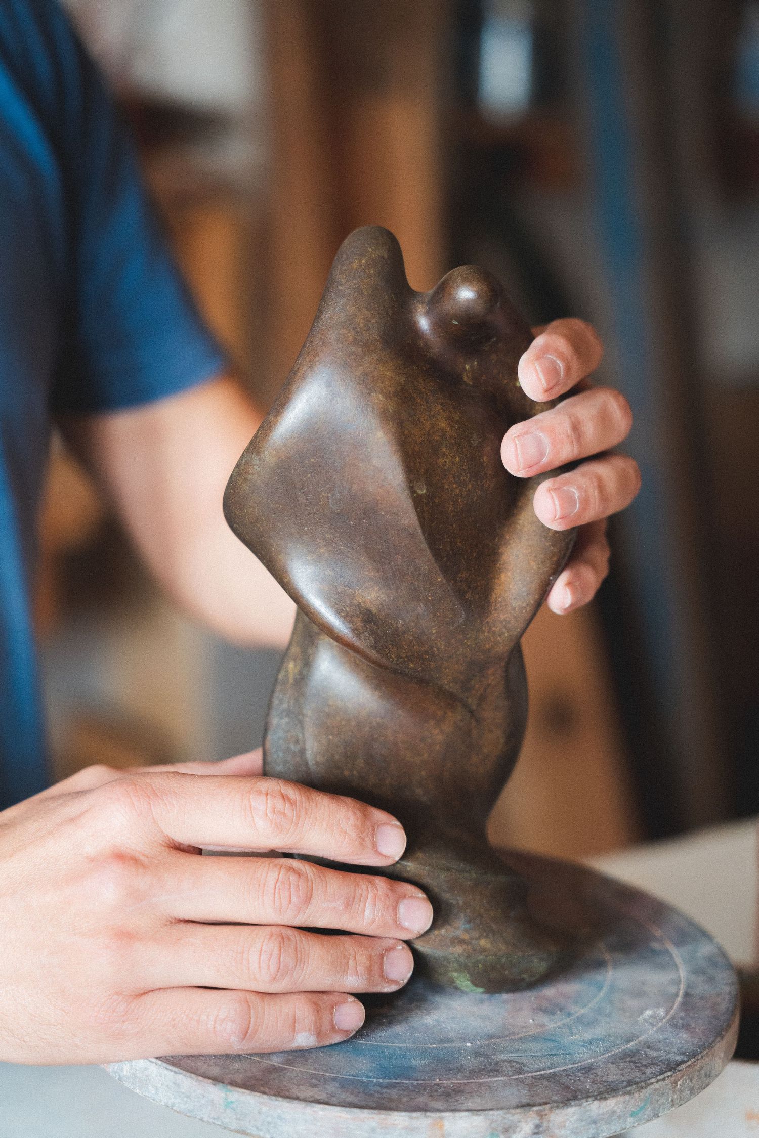 Artist's hands holding a smooth abstract bronze sculpture of a human torso on a rotating display stand.