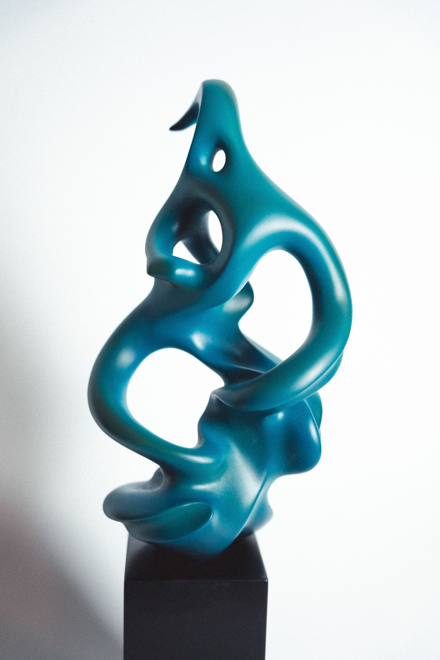 Abstract teal sculpture with fluid organic curves mounted on black pedestal against white background.
