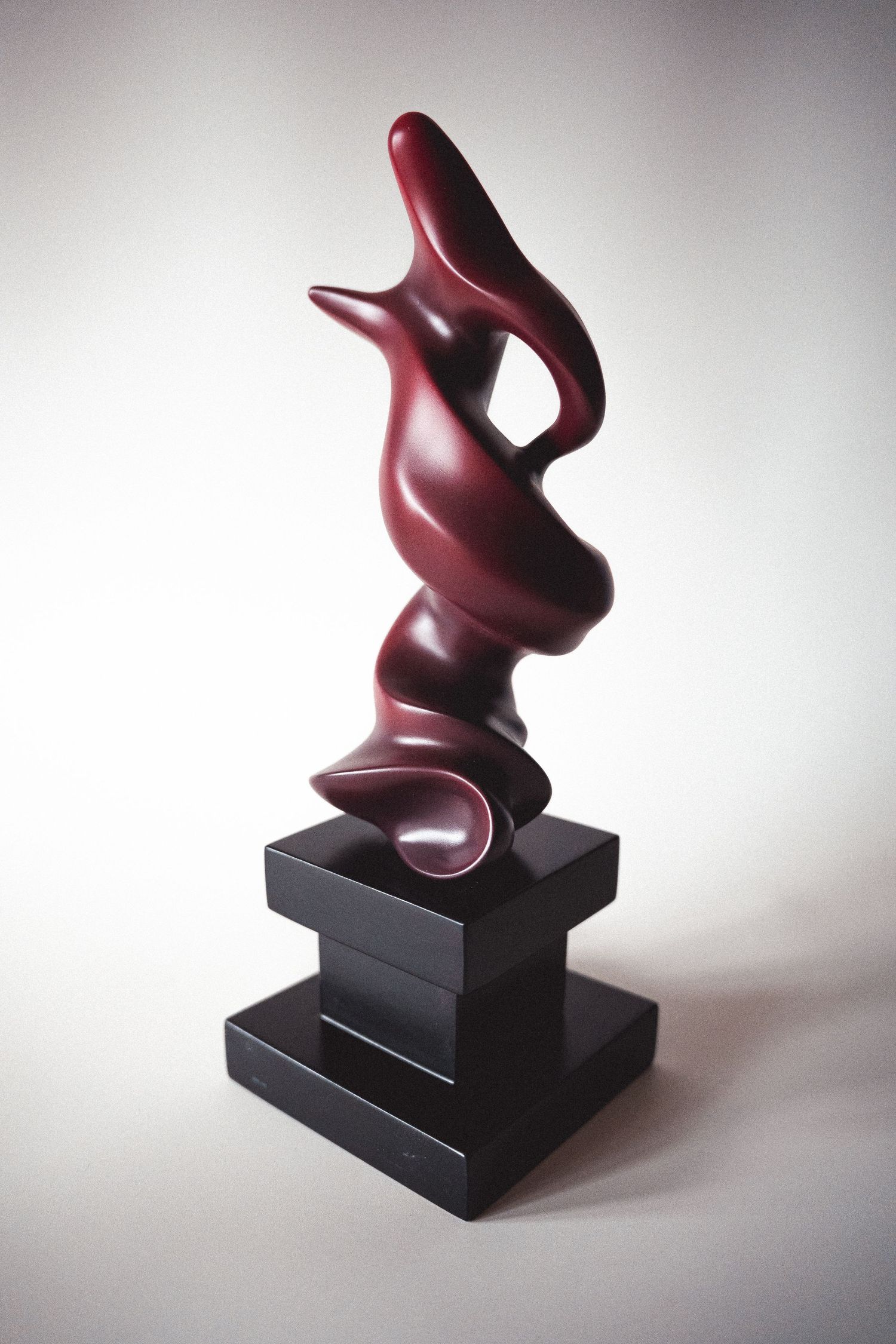 Abstract dark red sculpture with flowing organic form on a black tiered pedestal.