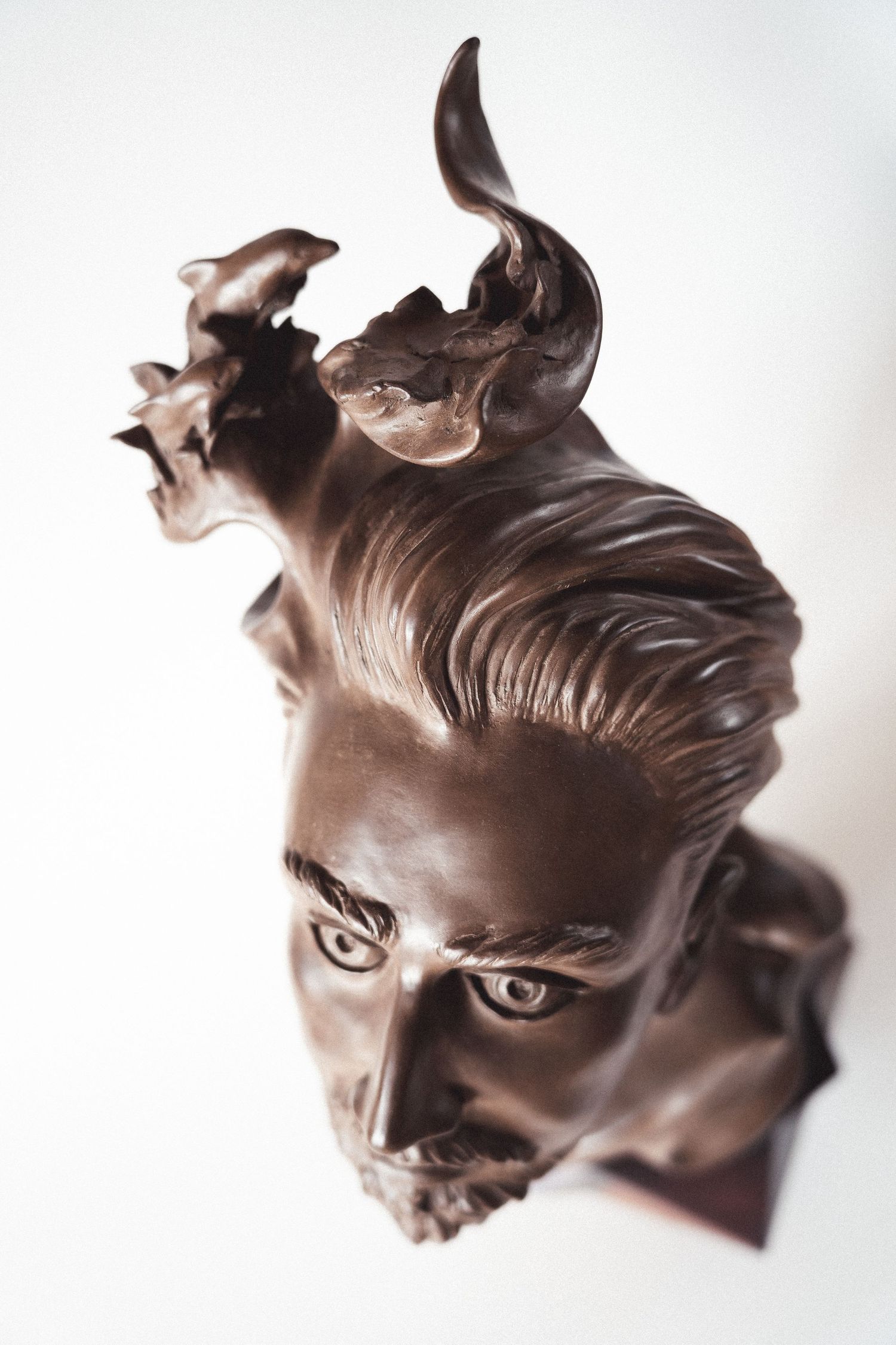 Bronze bust sculpture of a horned male figure viewed from above on white background.