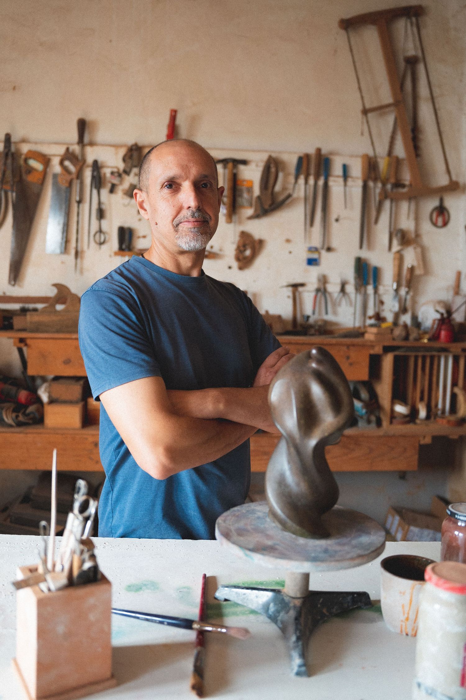 Confident male sculptor posing with arms crossed beside a dark abstract sculpture in workshop.