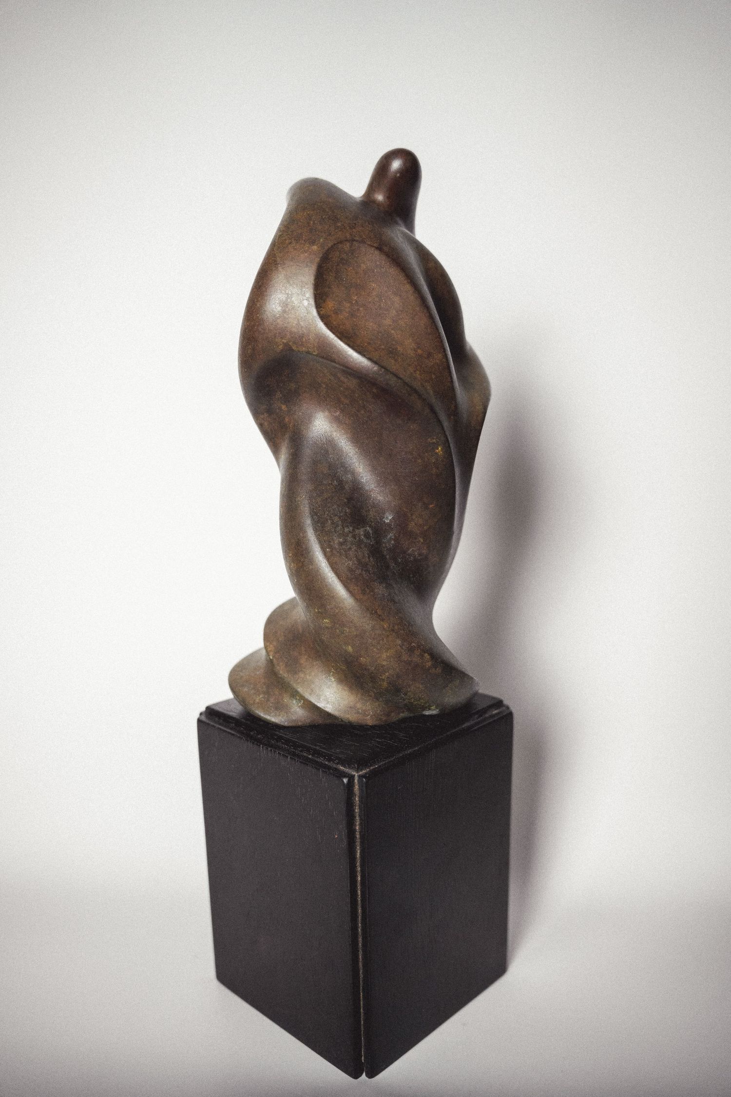 Dark bronze abstract figurative sculpture with sinuous twisting form displayed on a black pedestal.