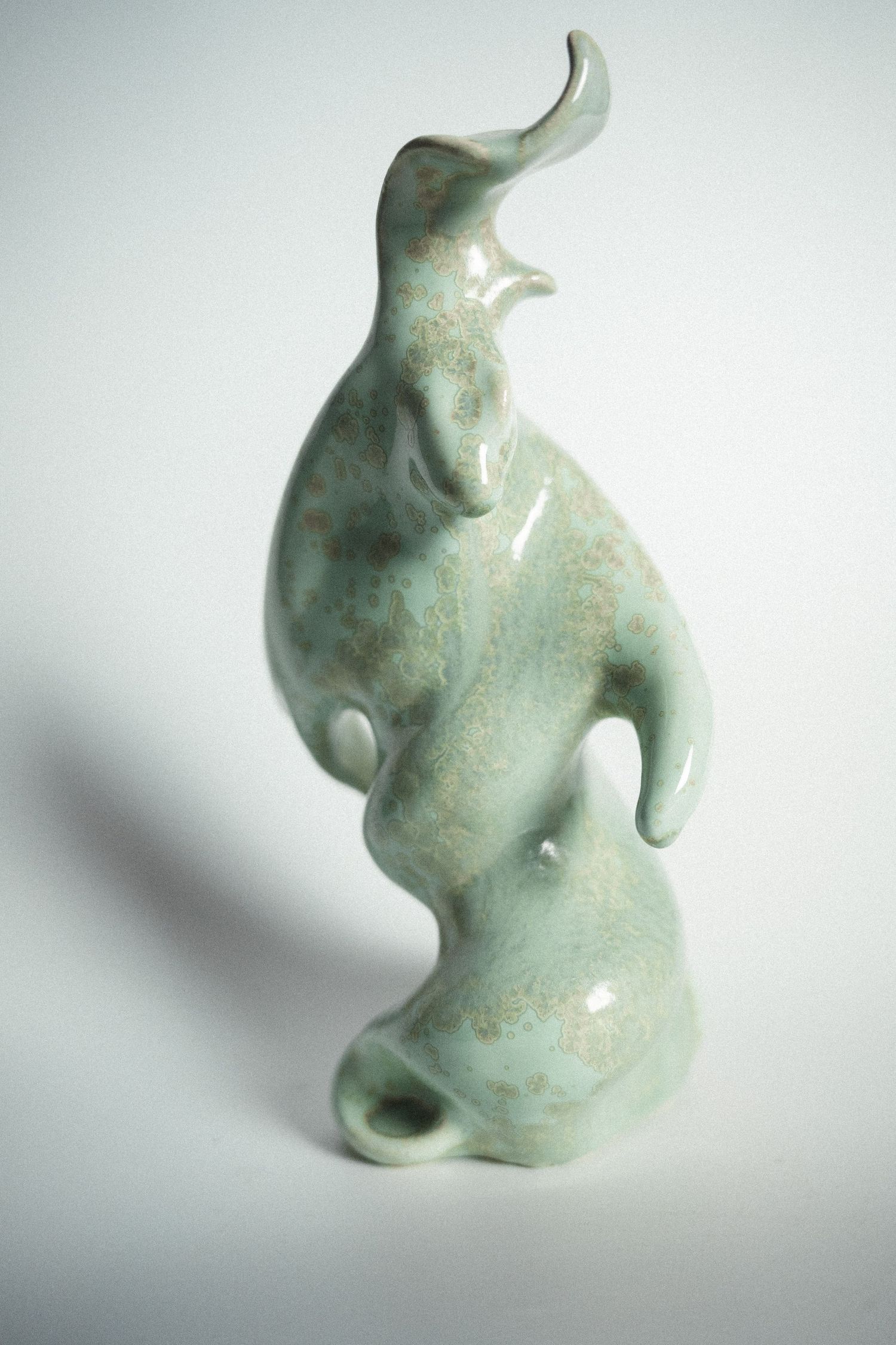 Abstract ceramic bird sculpture with celadon green glaze and flowing organic form on white background.