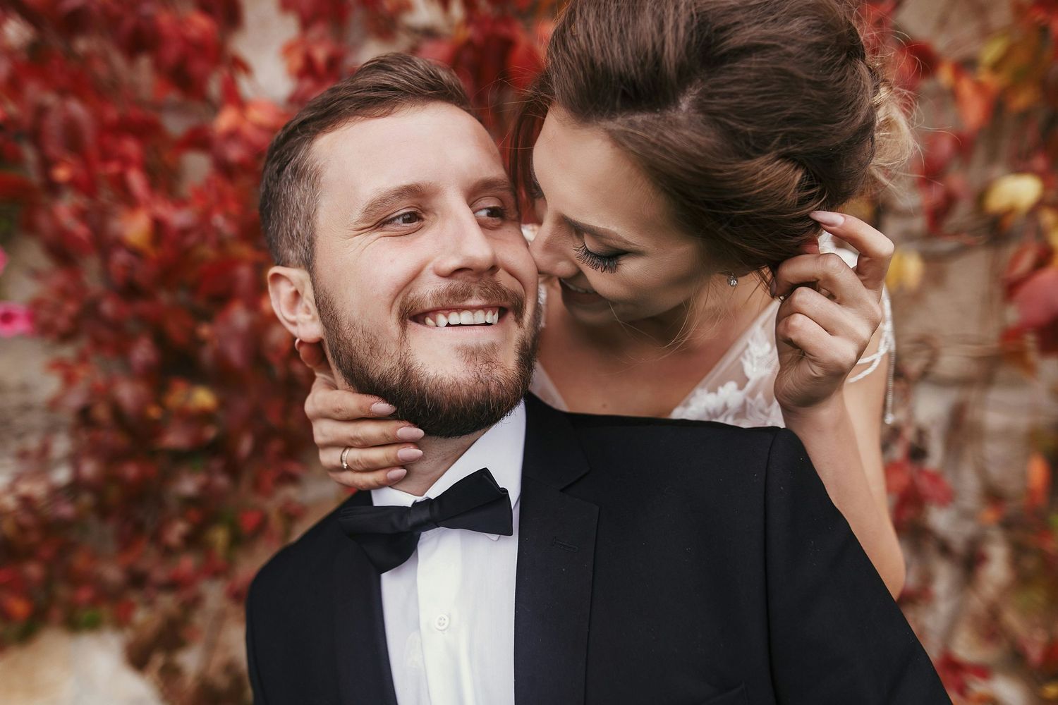 A smiling groom in a tuxedo embraces his bride against a backdrop of vibrant red autumn foliage.