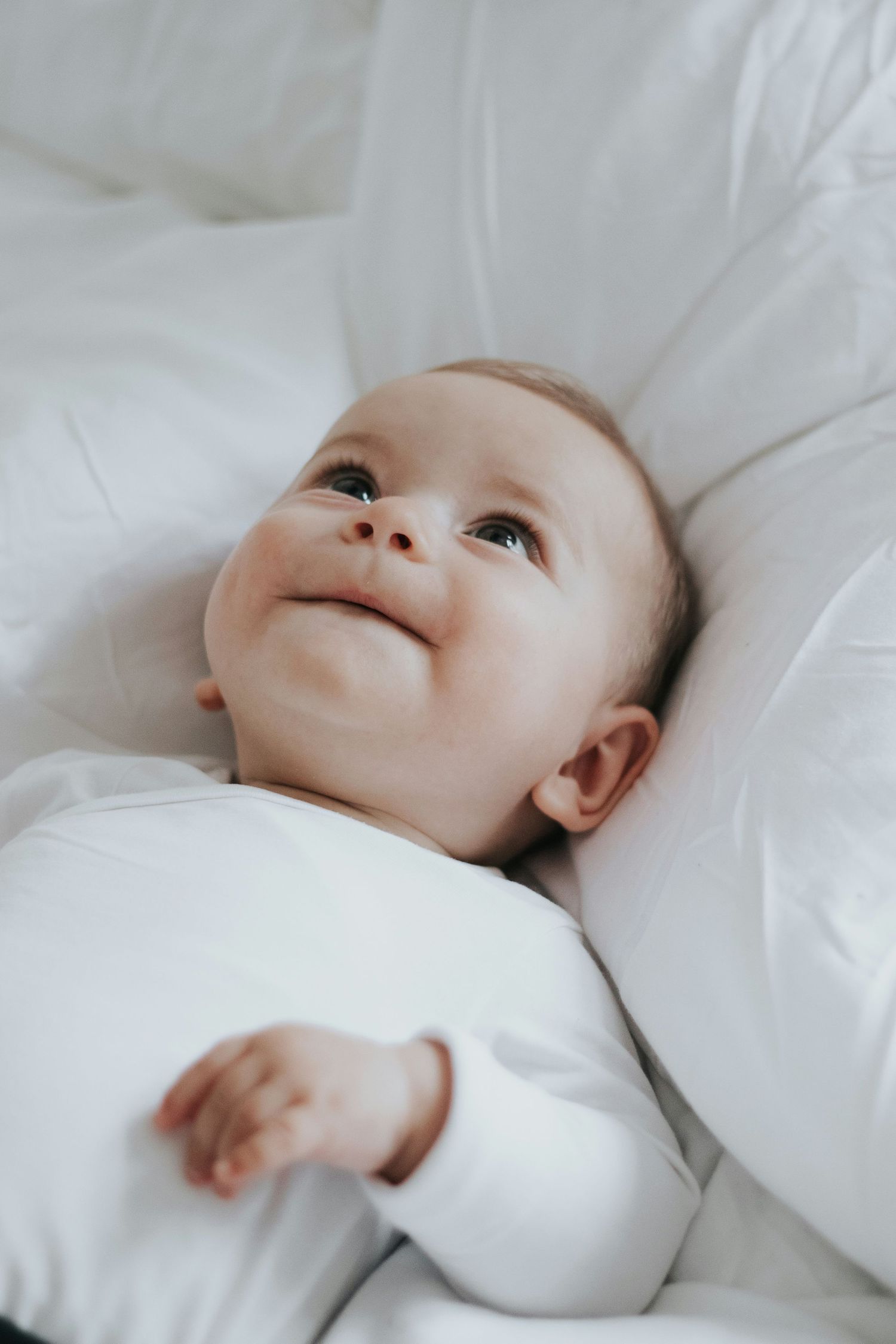A smiling baby in a white onesie lying on soft white bedding, gazing upward with a joyful expression.