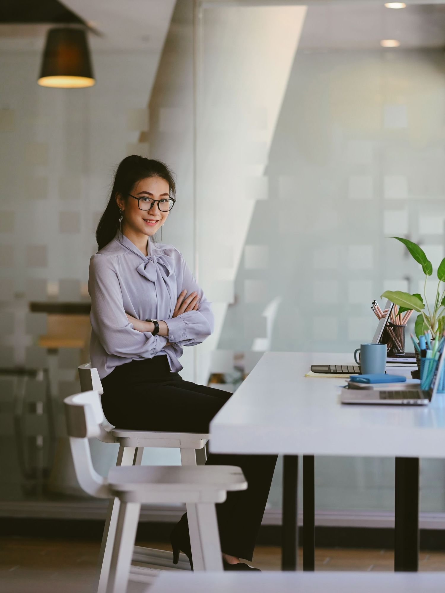 A confident young Asian businesswoman in glasses and a blue blouse sits on a desk with arms crossed in a modern office.