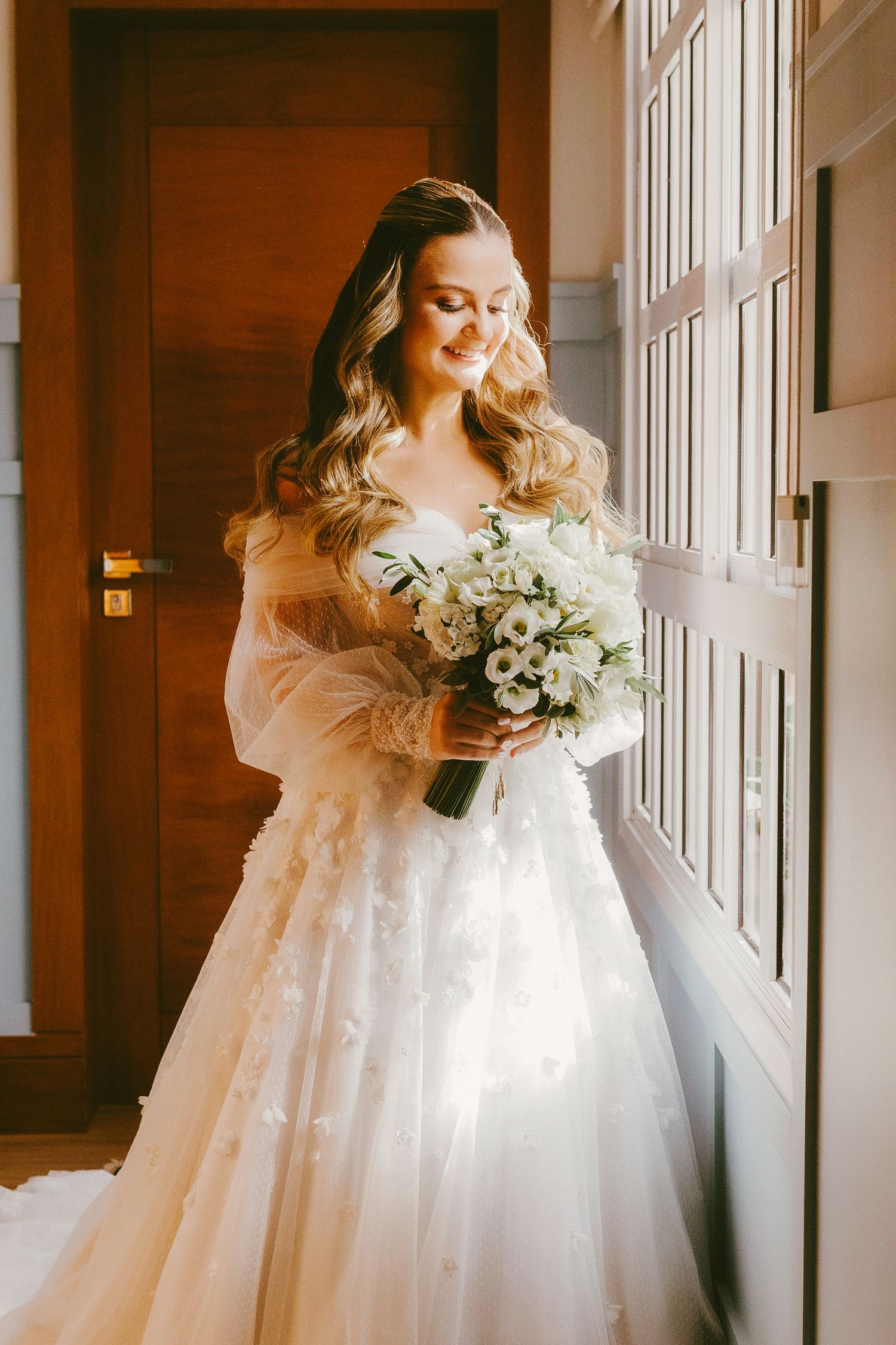 Bride in a white tulle ball gown with floral details holds a white bouquet near a sunlit window.
