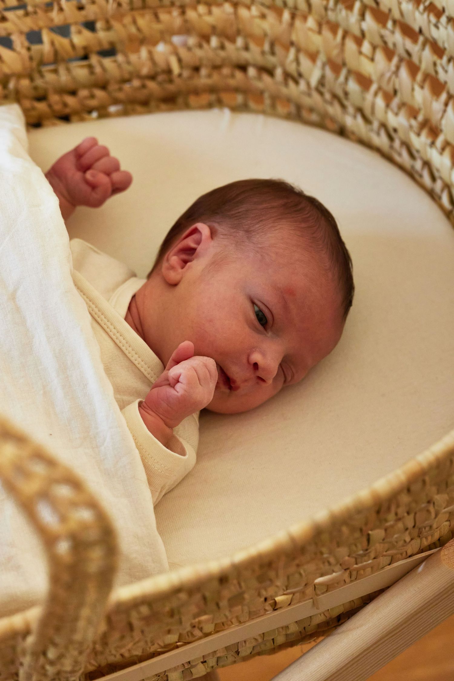 Newborn baby lying in a woven Moses basket, wearing cream outfit with hand near mouth.