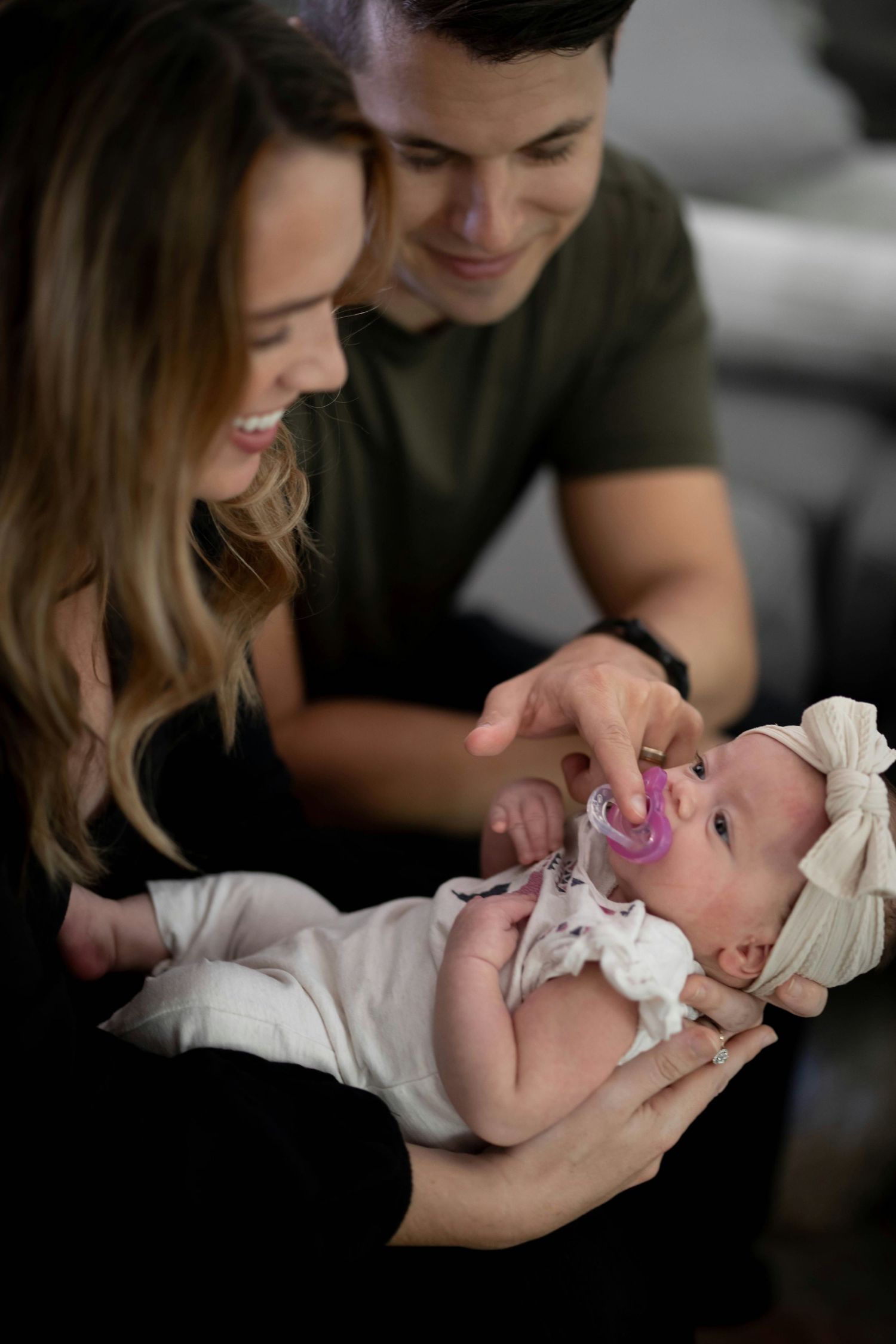 Smiling young parents admire their newborn baby girl with a pacifier and white bow headband.