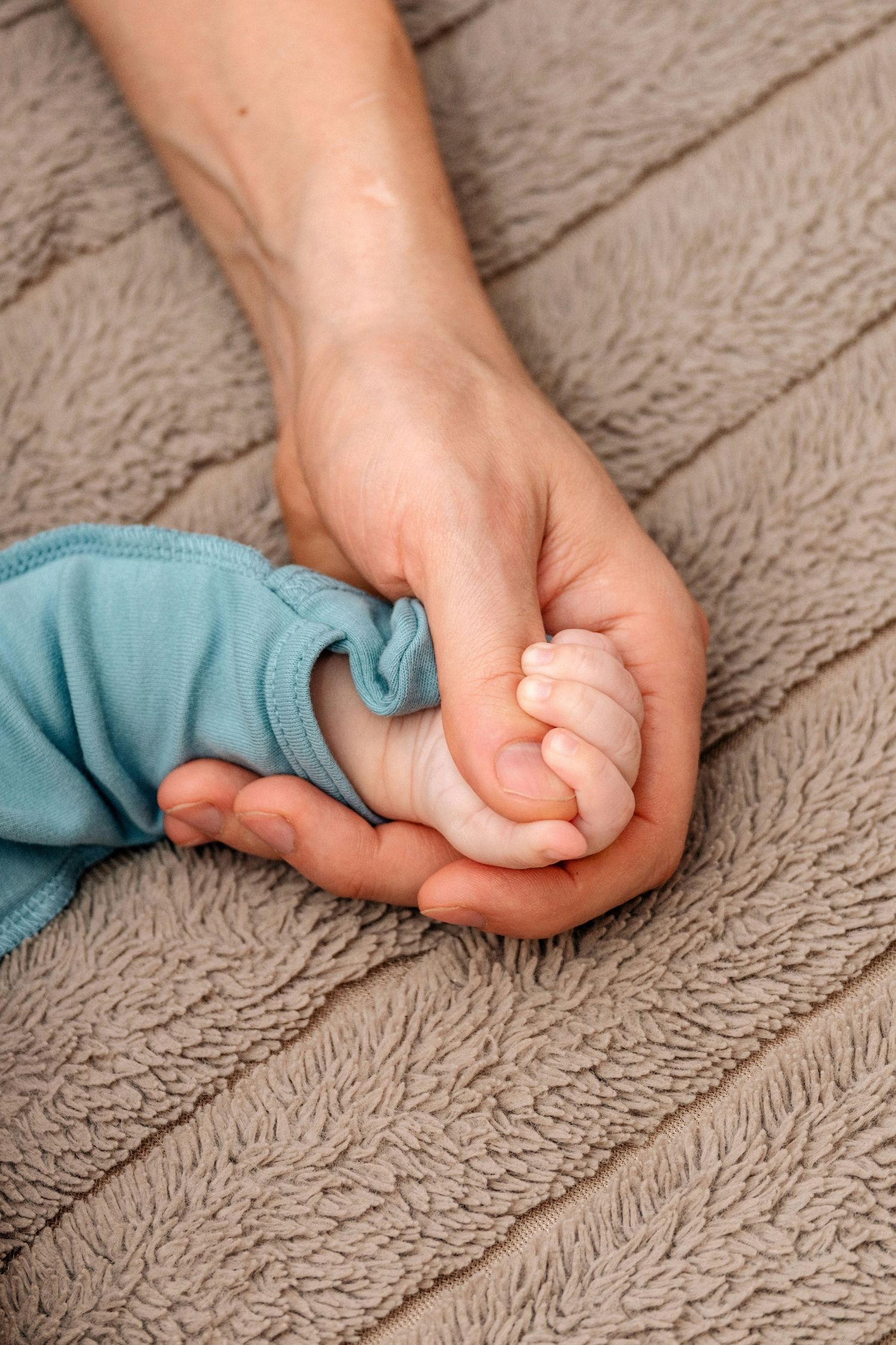Adult hand gently holding tiny newborn baby fist on brown textured towel.