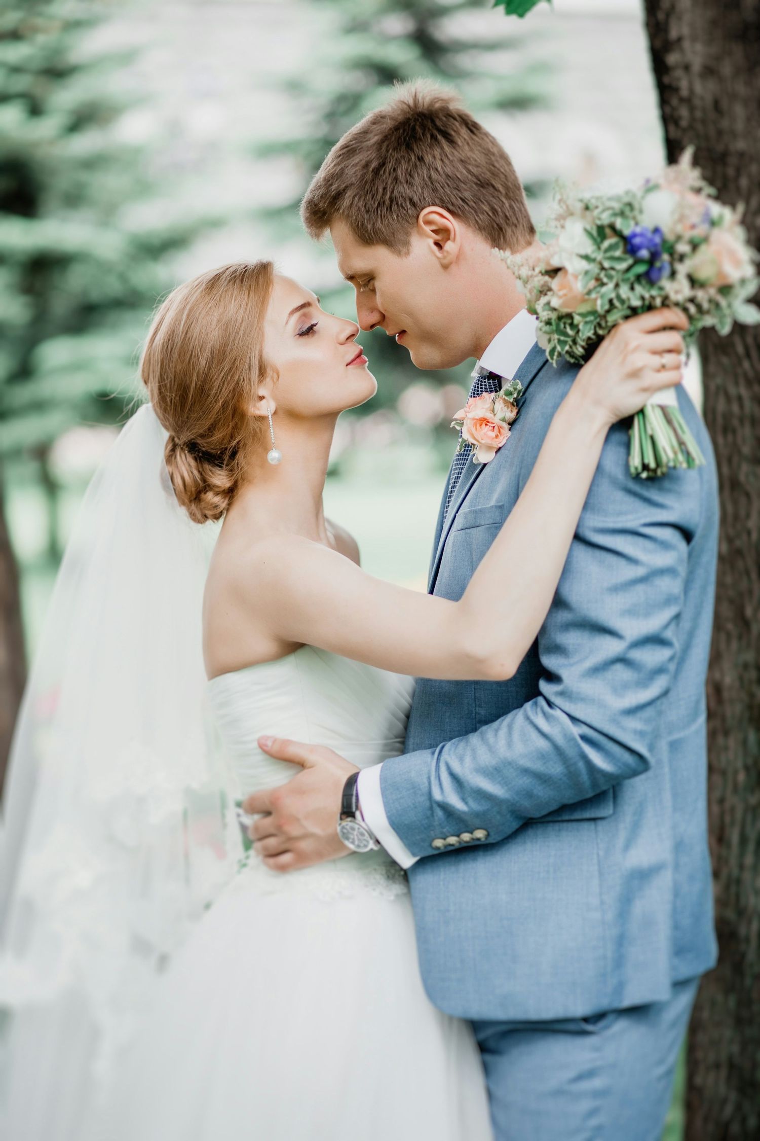 Wedding couple embraces near a tree outdoors, bride in white gown holding a colorful bouquet of flowers.
