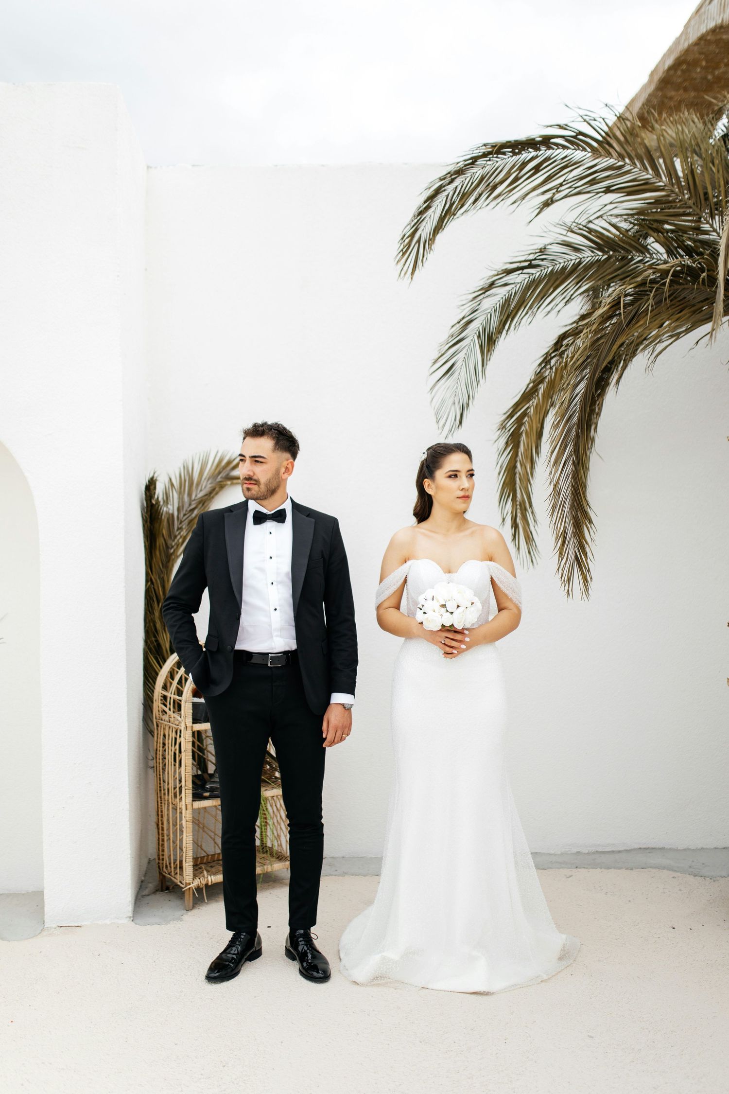 Bride and groom posing outdoors by white wall with palm tree, groom in black tuxedo.