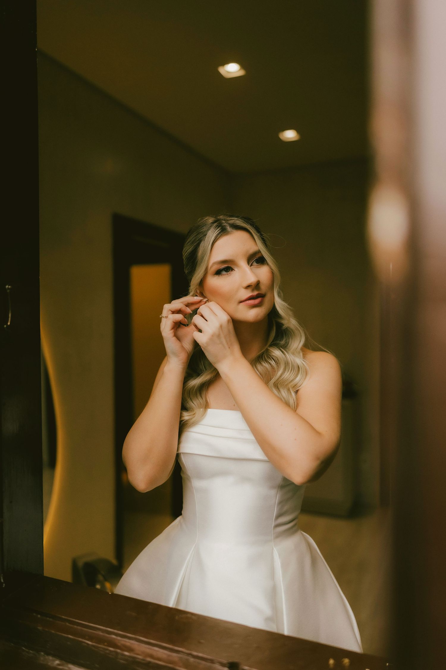 Bride in white strapless gown putting on earrings while getting ready, reflected in mirror with warm lighting.