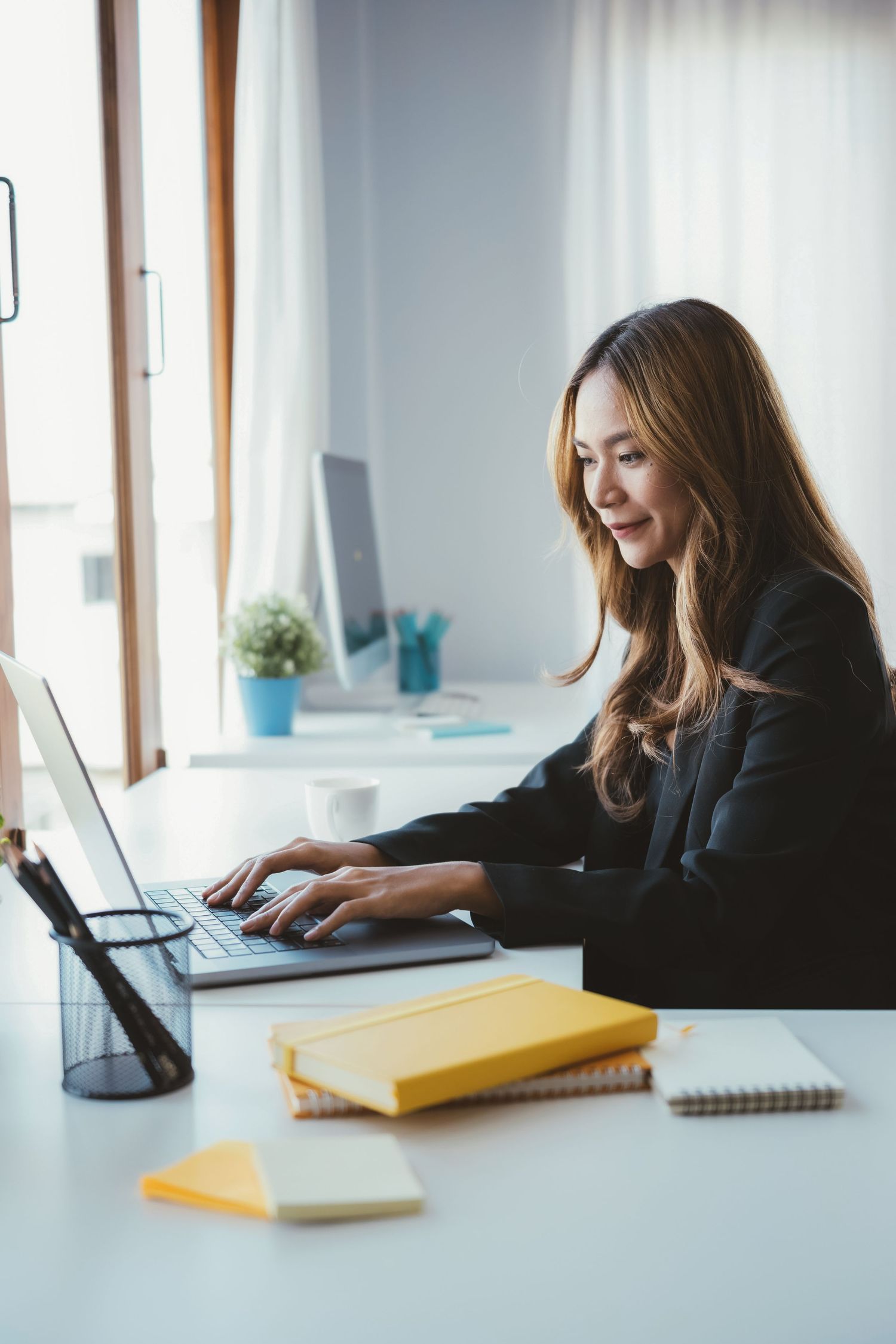 Smiling Asian businesswoman in black blazer typing on laptop at organized white office desk.