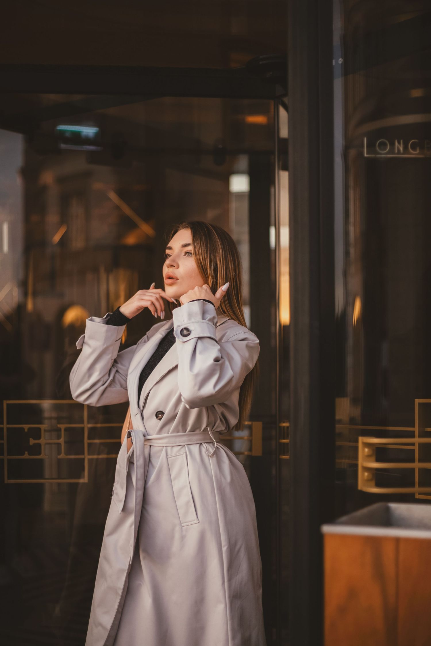Elegant woman in light trench coat posing outside a city storefront at night.