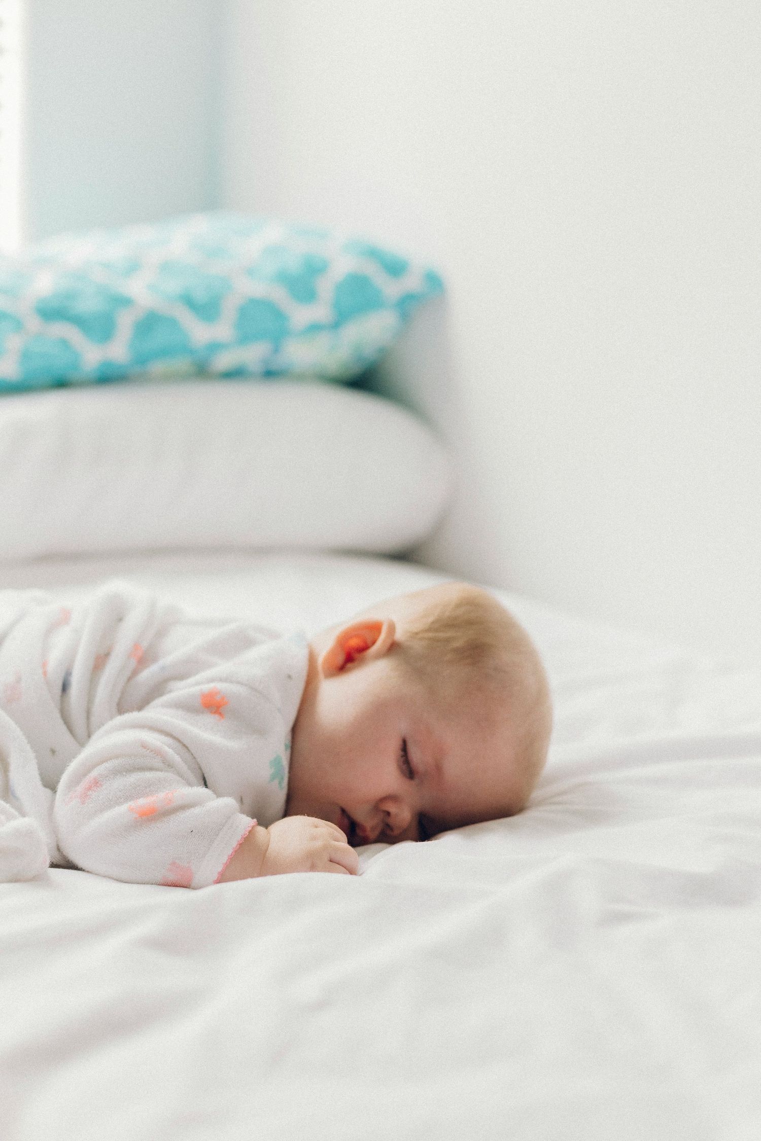 Sleeping baby in a white floral onesie lying face-down on white bedding with teal pillows in background.