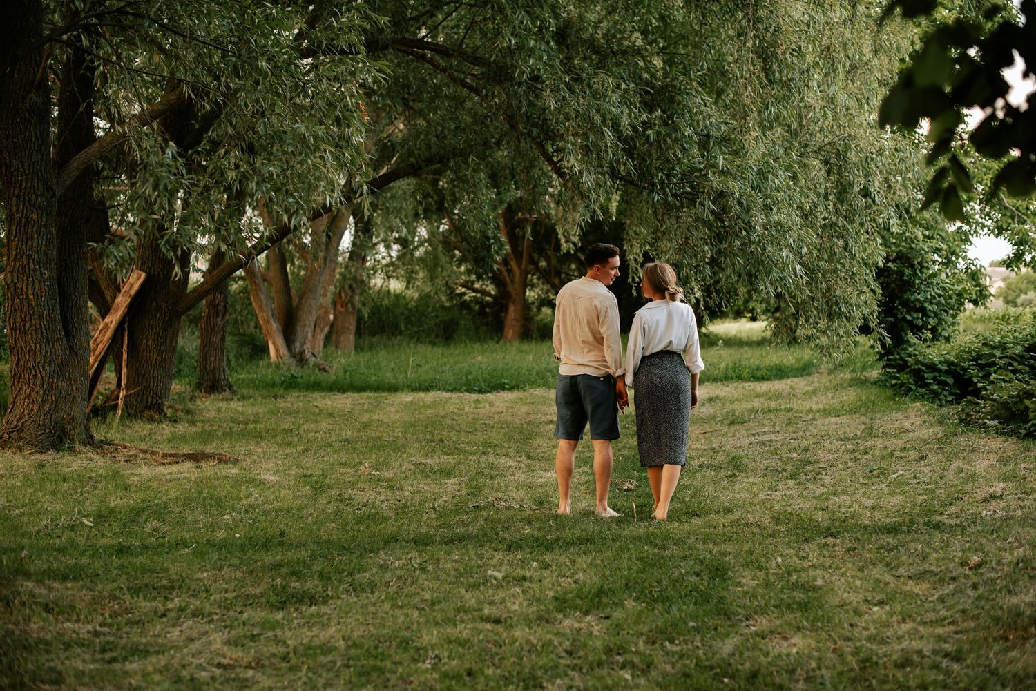 Couple walking hand in hand through a serene green park surrounded by willow trees.