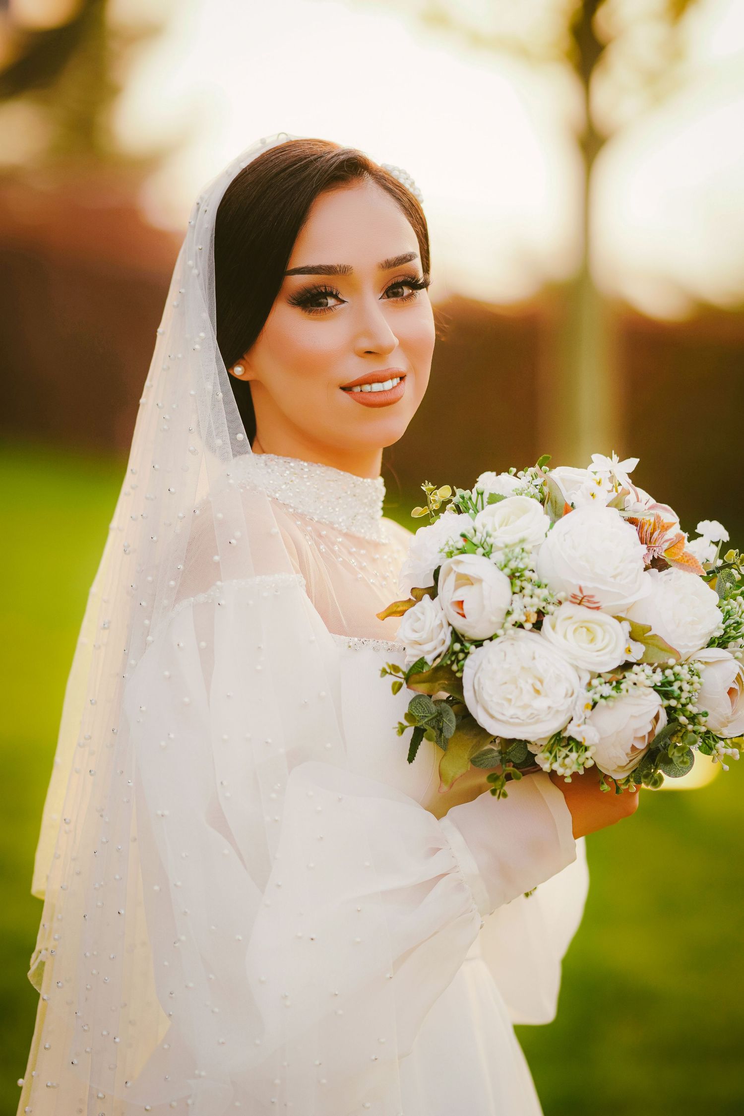 Elegant bride in white gown and veil holding a bouquet of white peonies, smiling outdoors in golden sunlight.