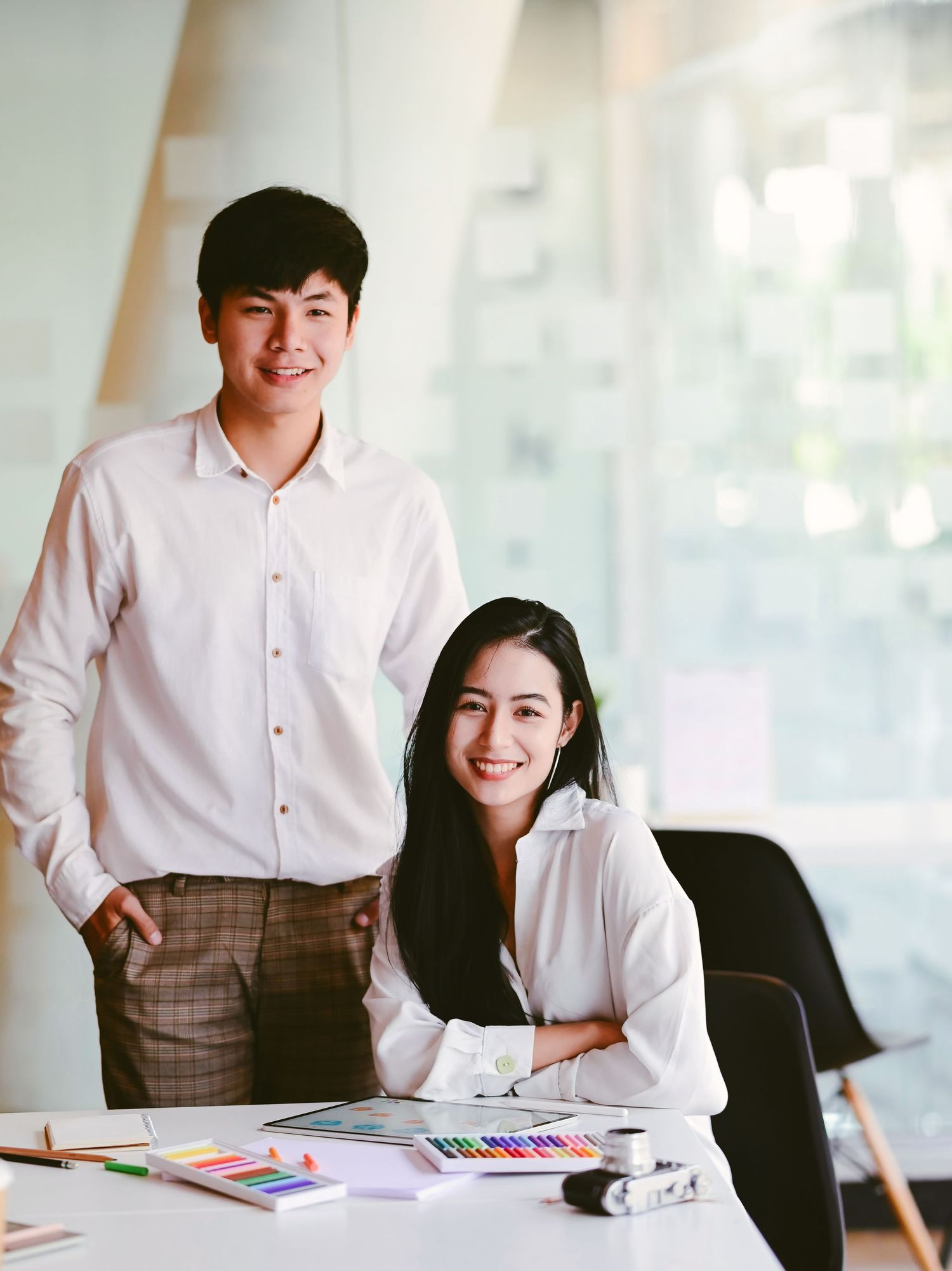Two smiling young Asian professionals pose together at a creative workspace with colorful design materials on the desk.