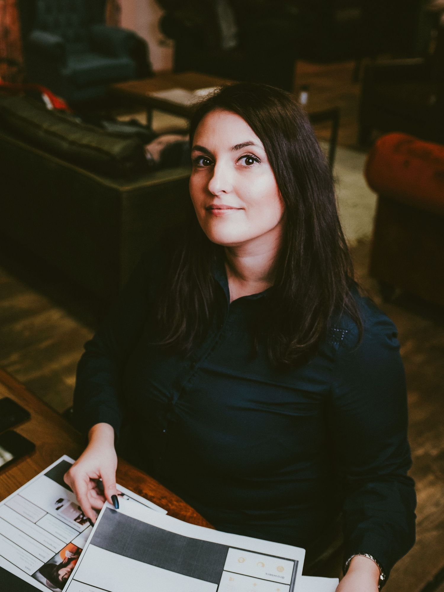 Young woman with dark hair sitting at a table in a cafe, holding a magazine and smiling.