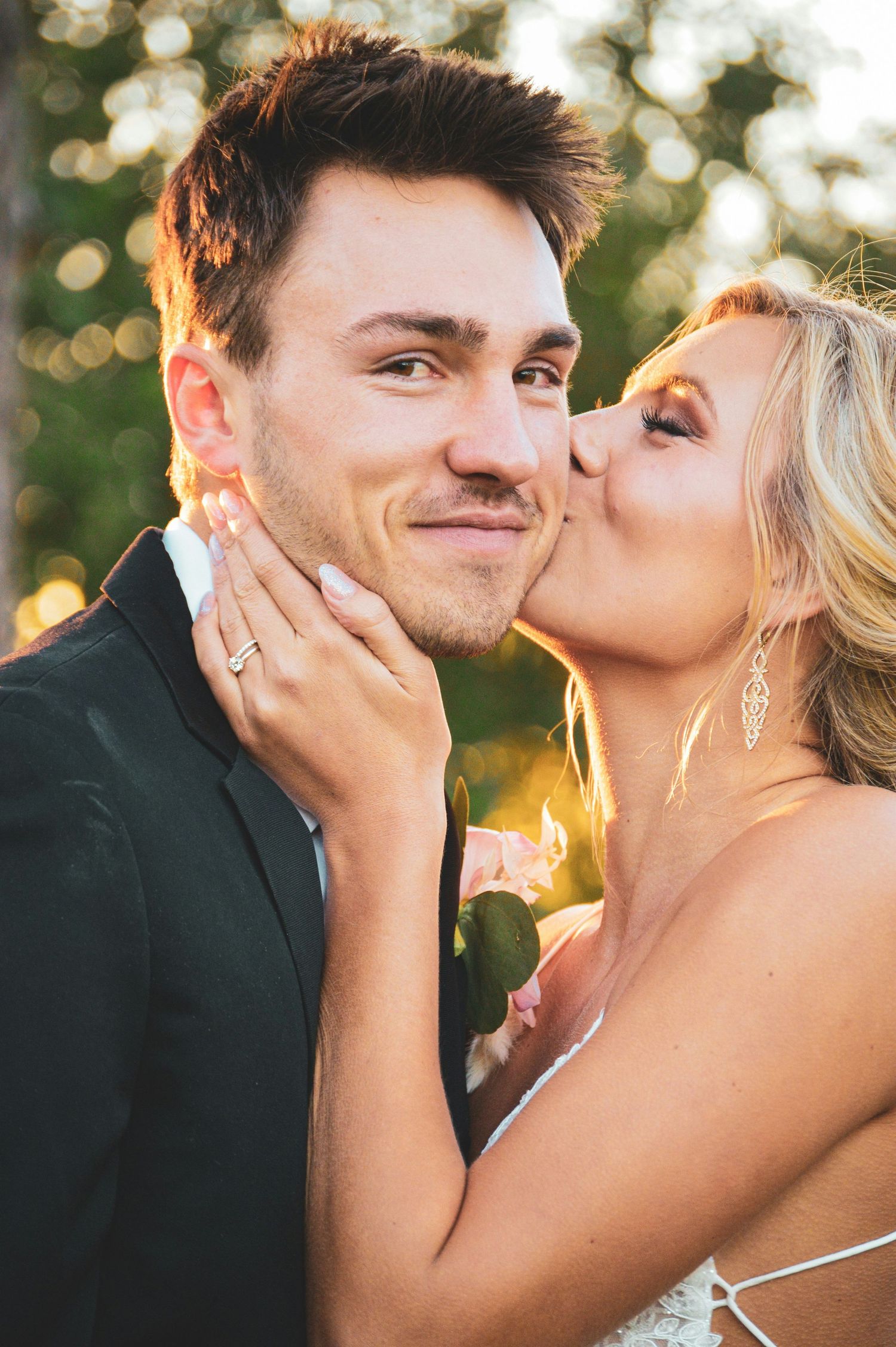 Blonde bride in white gown kisses groom on cheek during golden hour outdoor wedding portrait session.