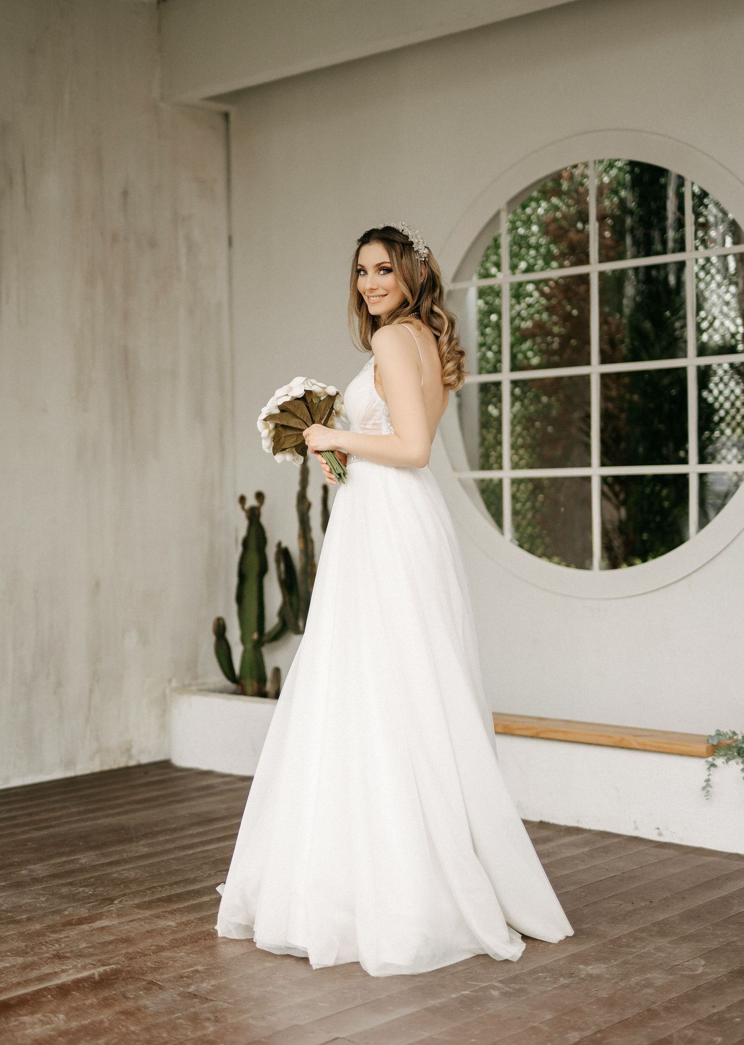 Smiling bride in flowing A-line wedding dress holding bouquet near round window with cactus.
