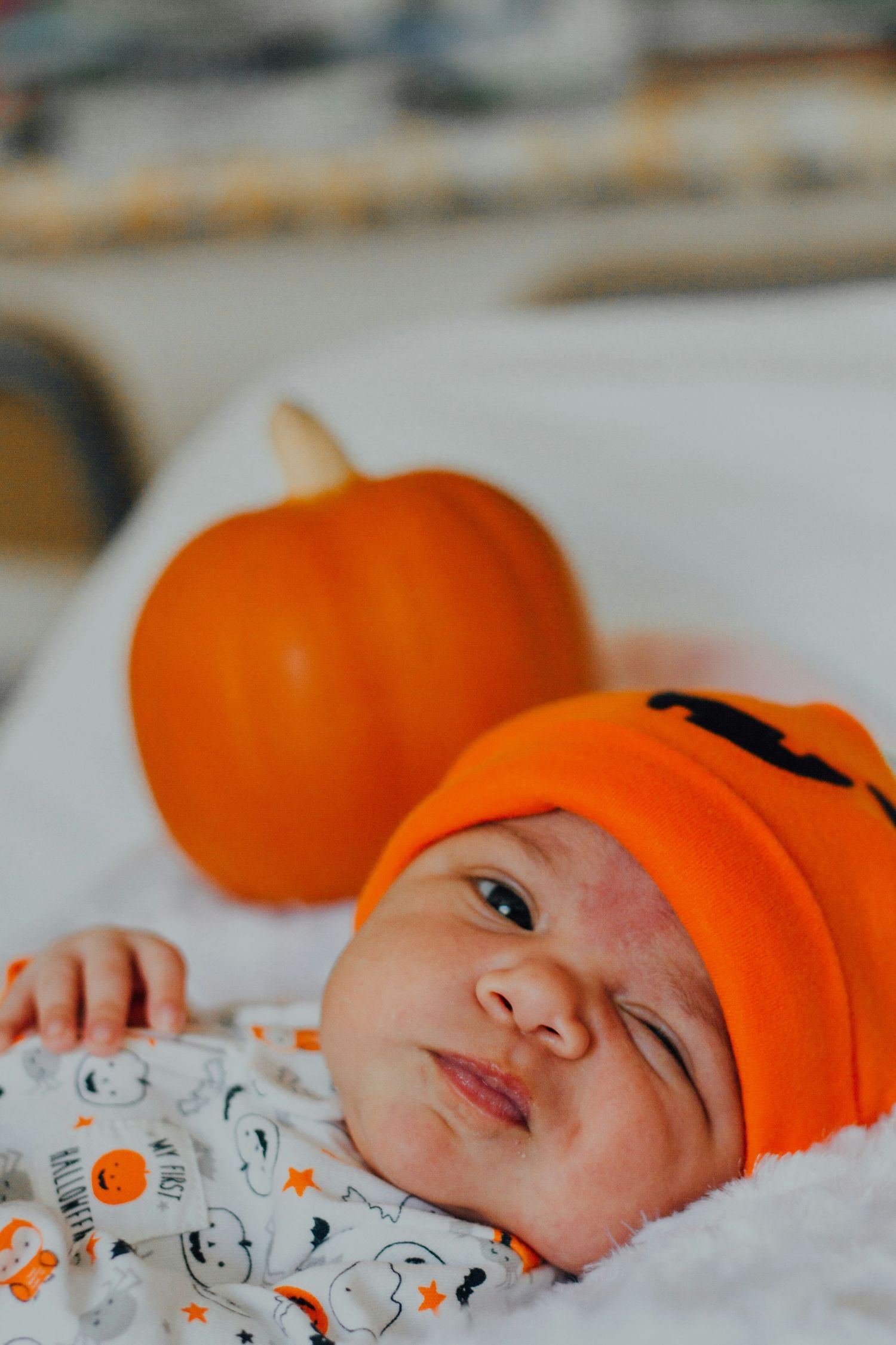 Newborn baby wearing an orange pumpkin hat lying next to a small orange pumpkin.