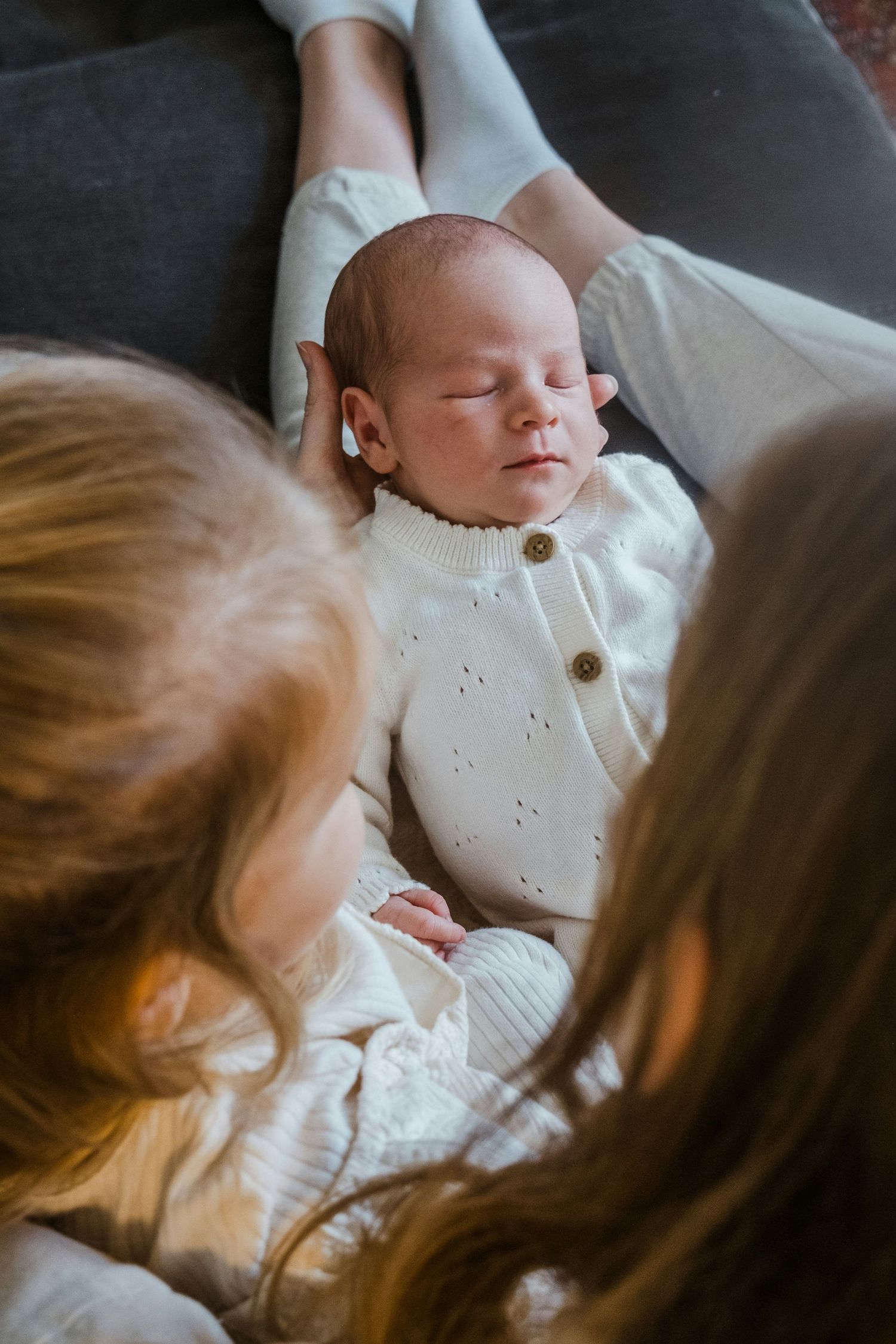 Newborn baby sleeping peacefully in white knit outfit while being held by mother and sibling.