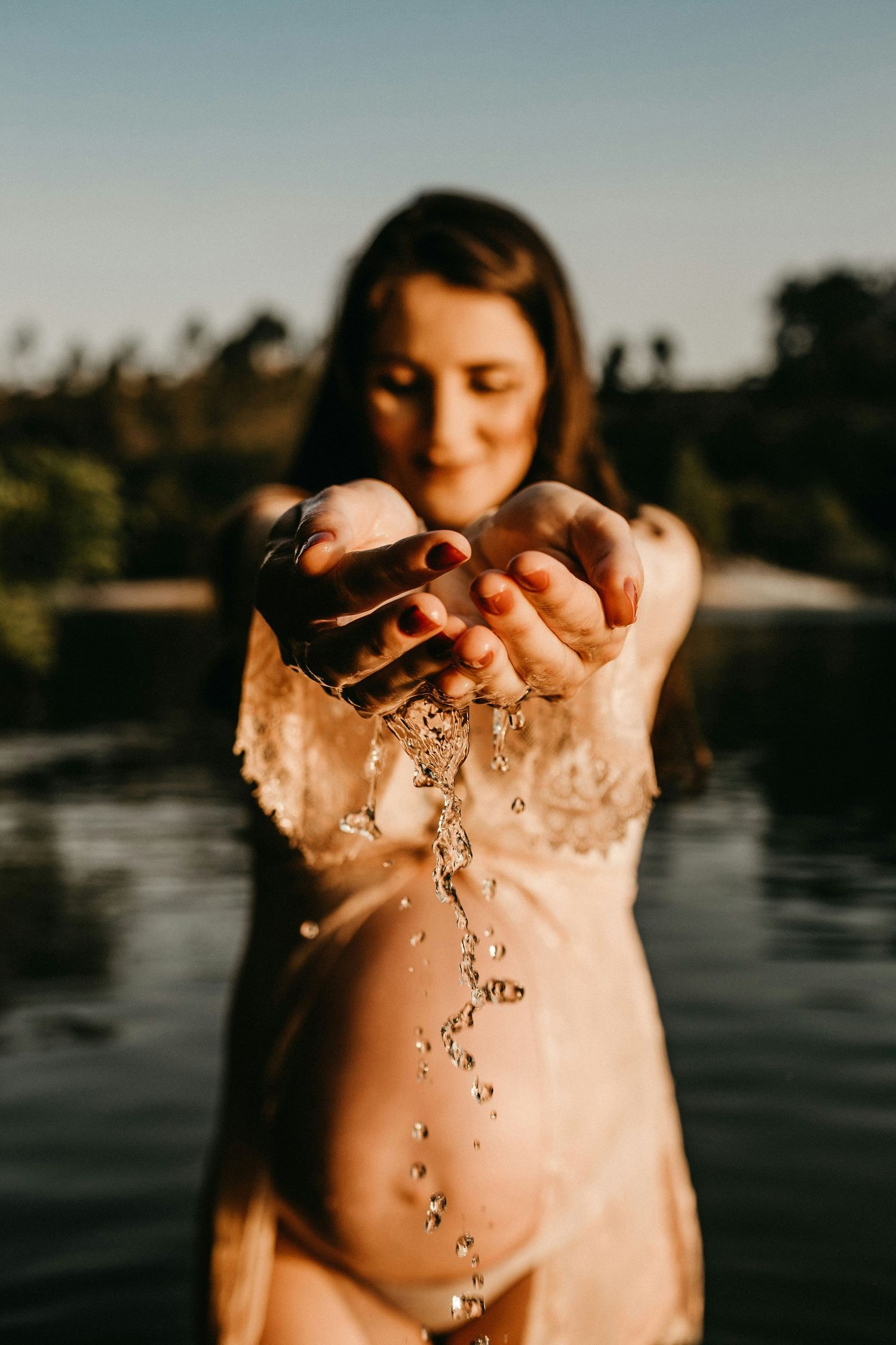 Pregnant woman standing in water pouring water from cupped hands at sunset.