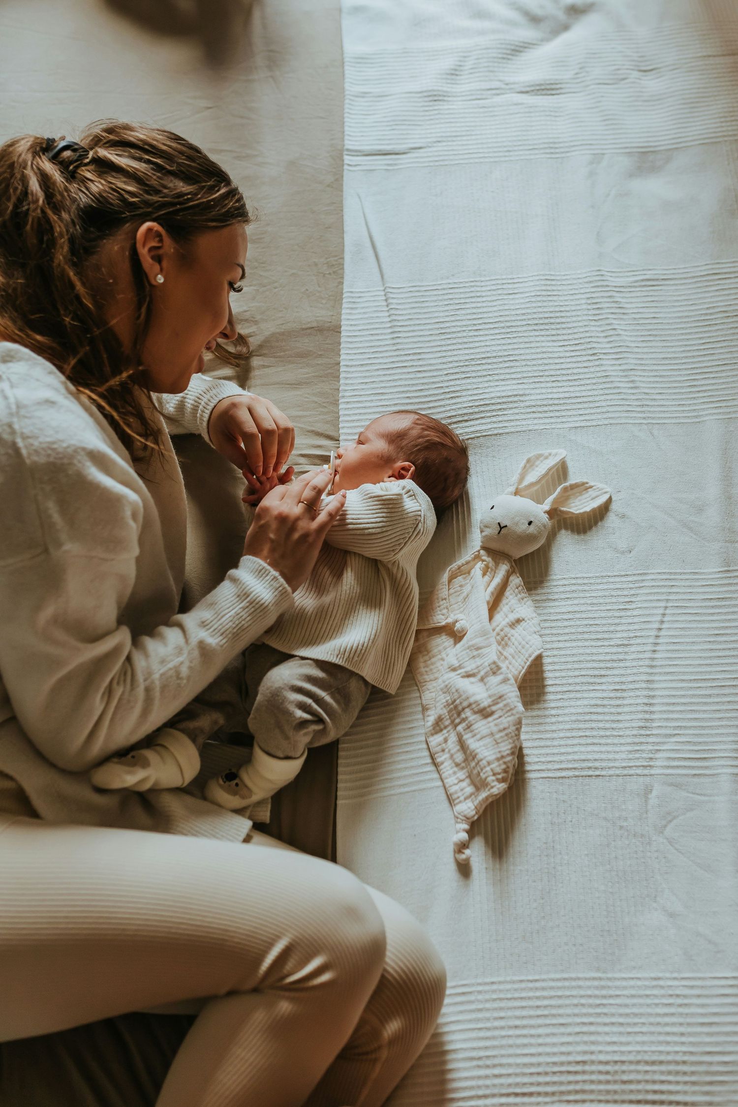 Mother in white outfit looking down at newborn baby lying on bed beside a stuffed bunny toy.