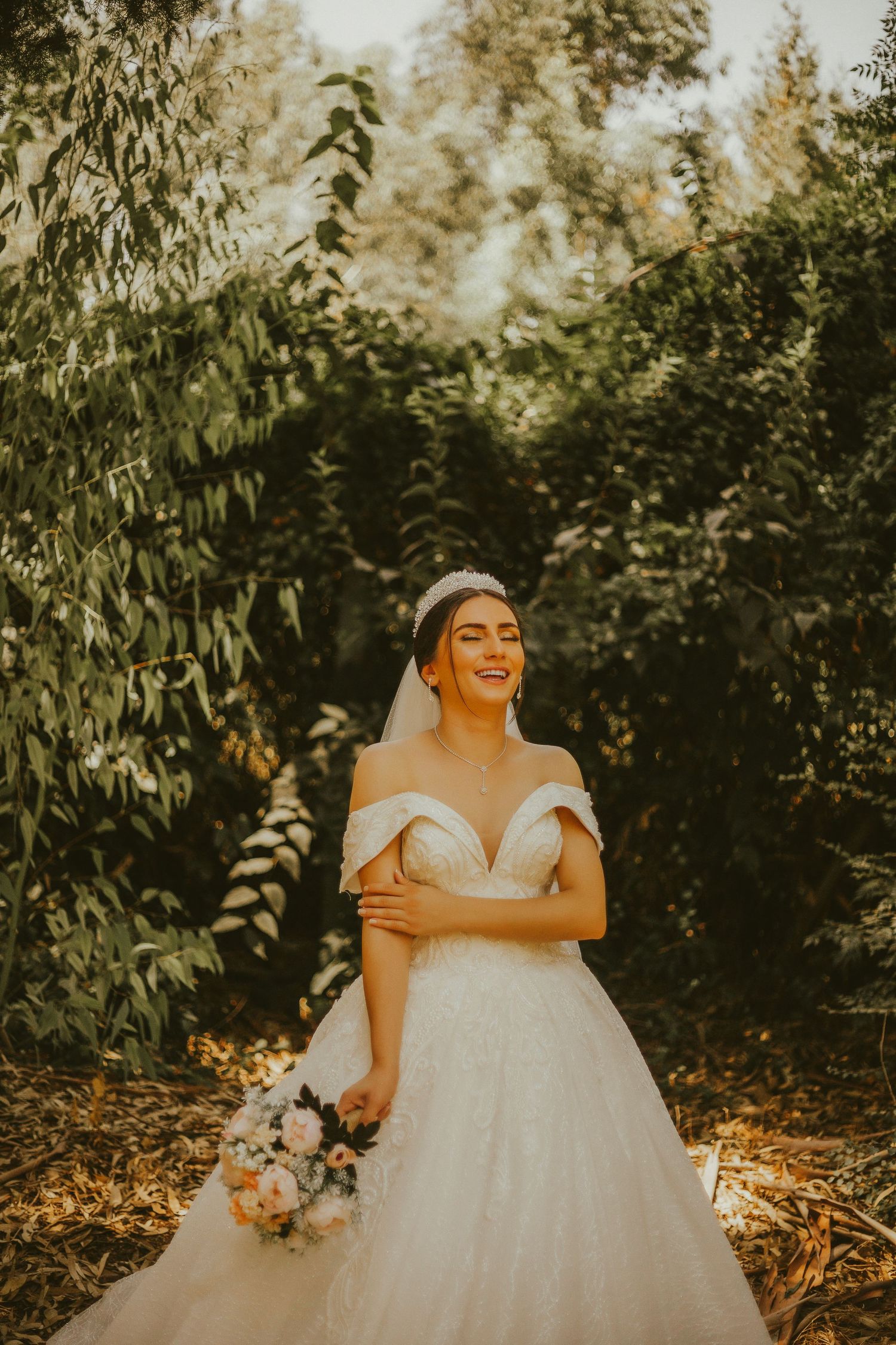 Smiling bride in off-shoulder white gown and tiara holds bouquet outdoors among lush green trees.