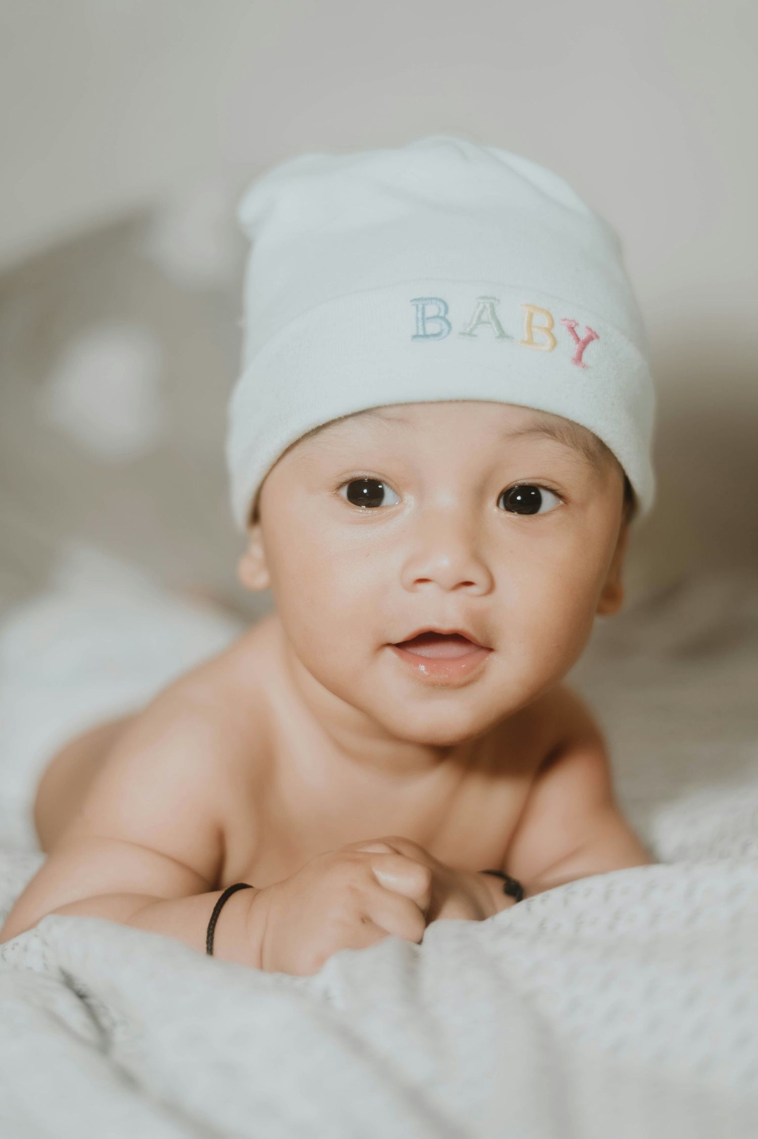 Cute baby wearing white hat embroidered with BABY doing tummy time on soft blanket.