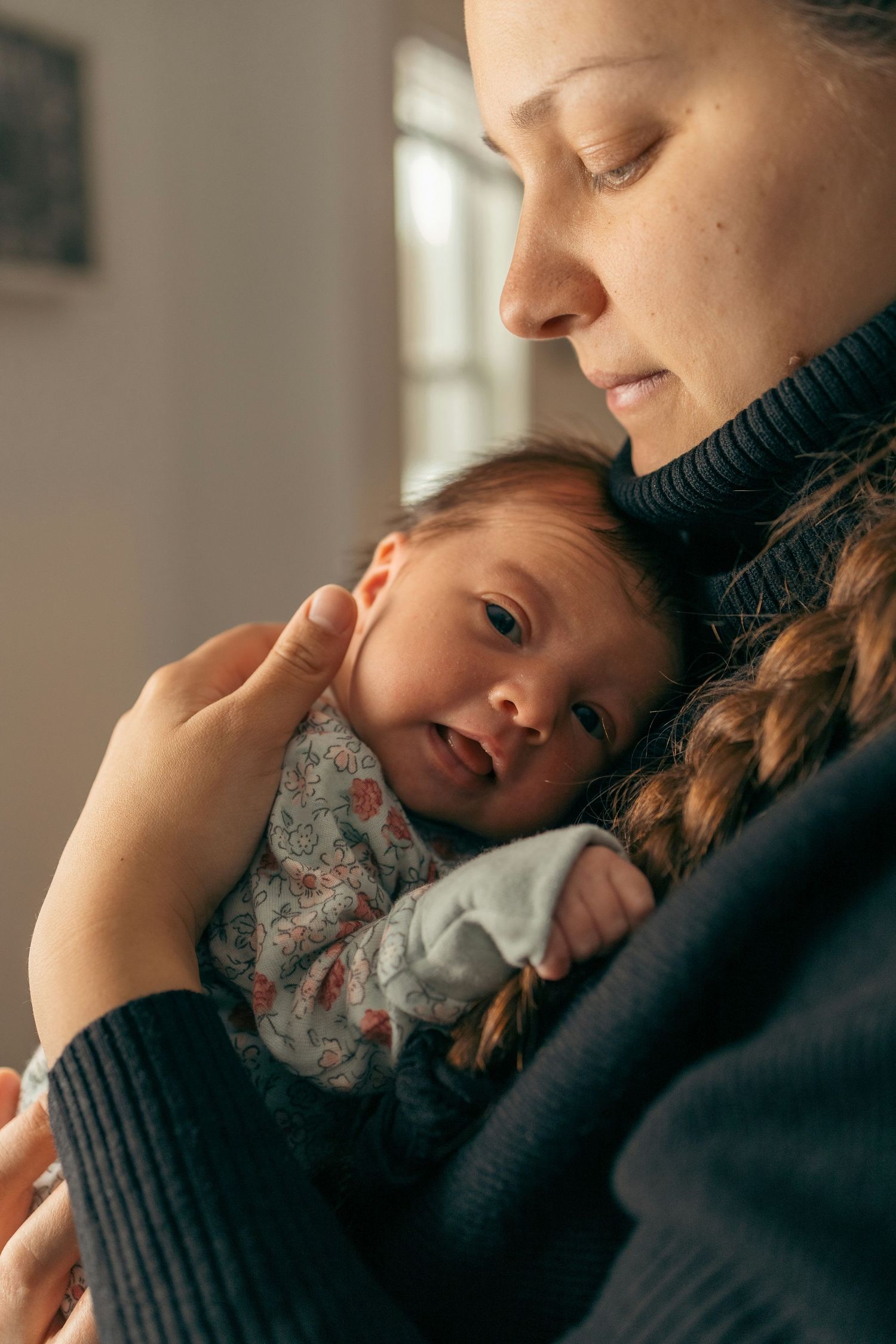 Mother with braided hair holding newborn baby close to her chest near a softly lit window.