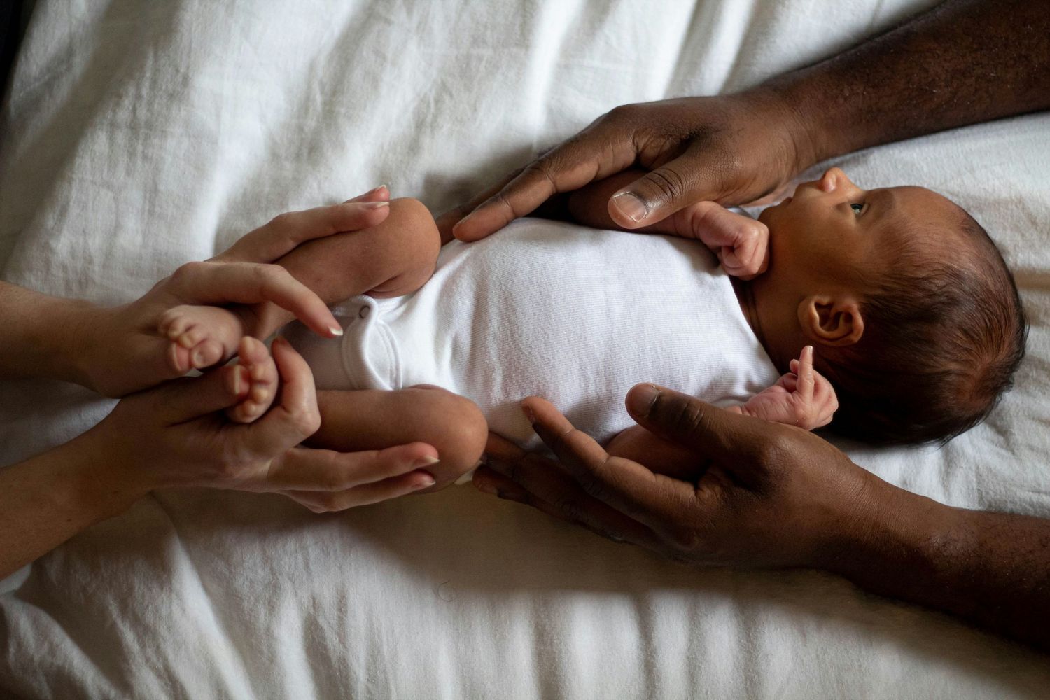 Parents hands cradling tiny newborn baby dressed in white on white sheet.