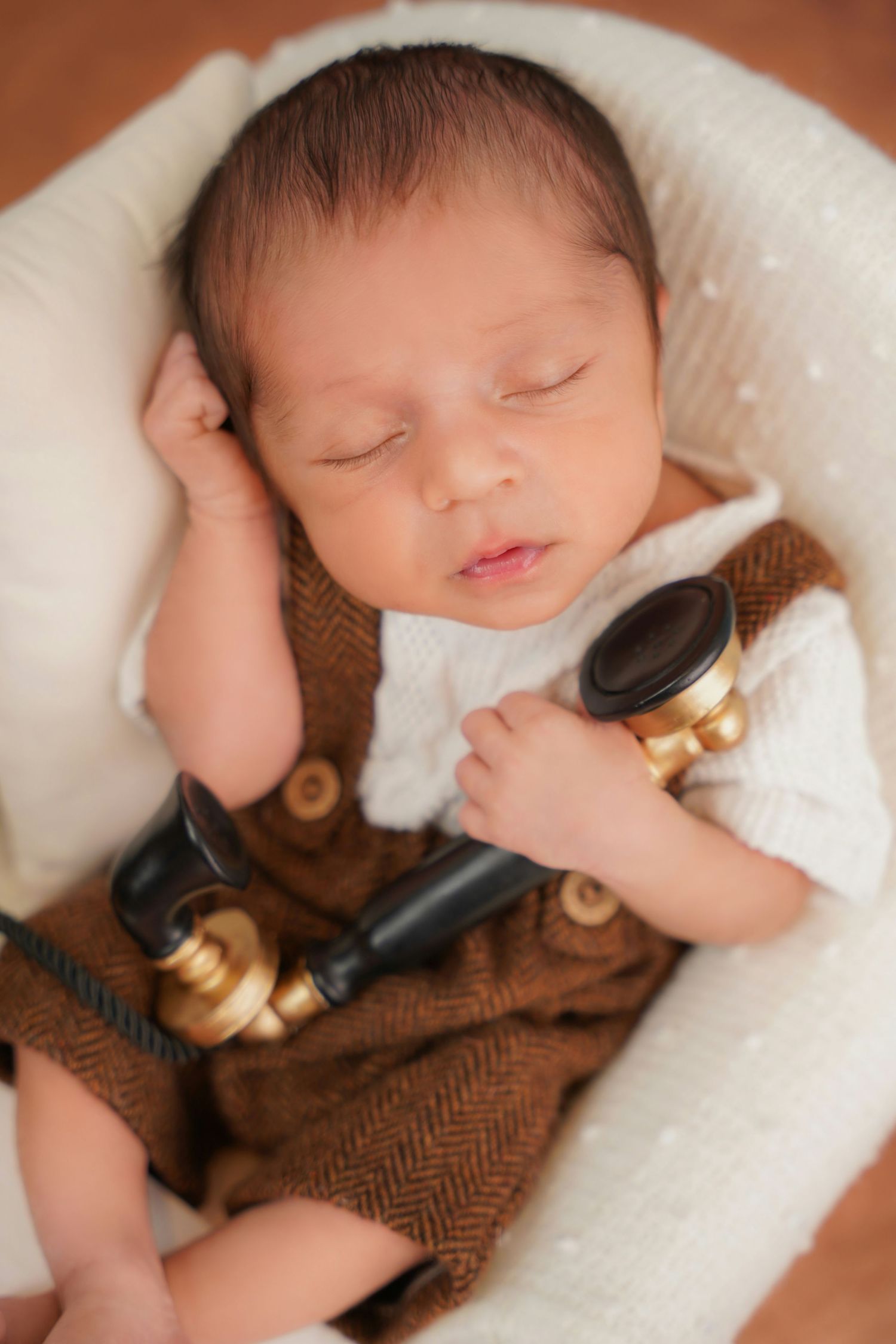 Sleeping newborn in brown herringbone outfit holding vintage telephone prop in white basket.
