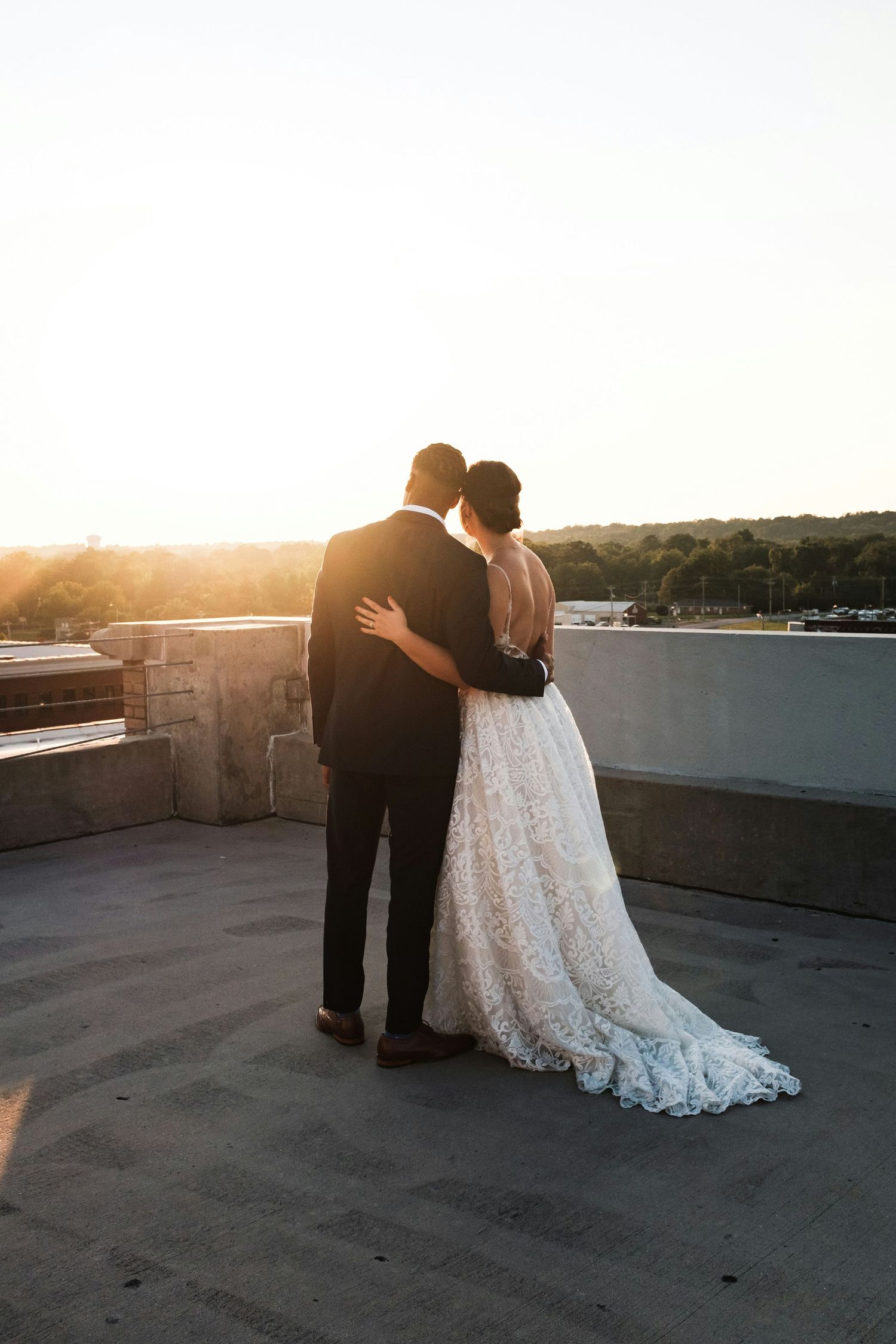 Bride and groom stand arm in arm on rooftop watching stunning sunset, bride in lace gown with long train.