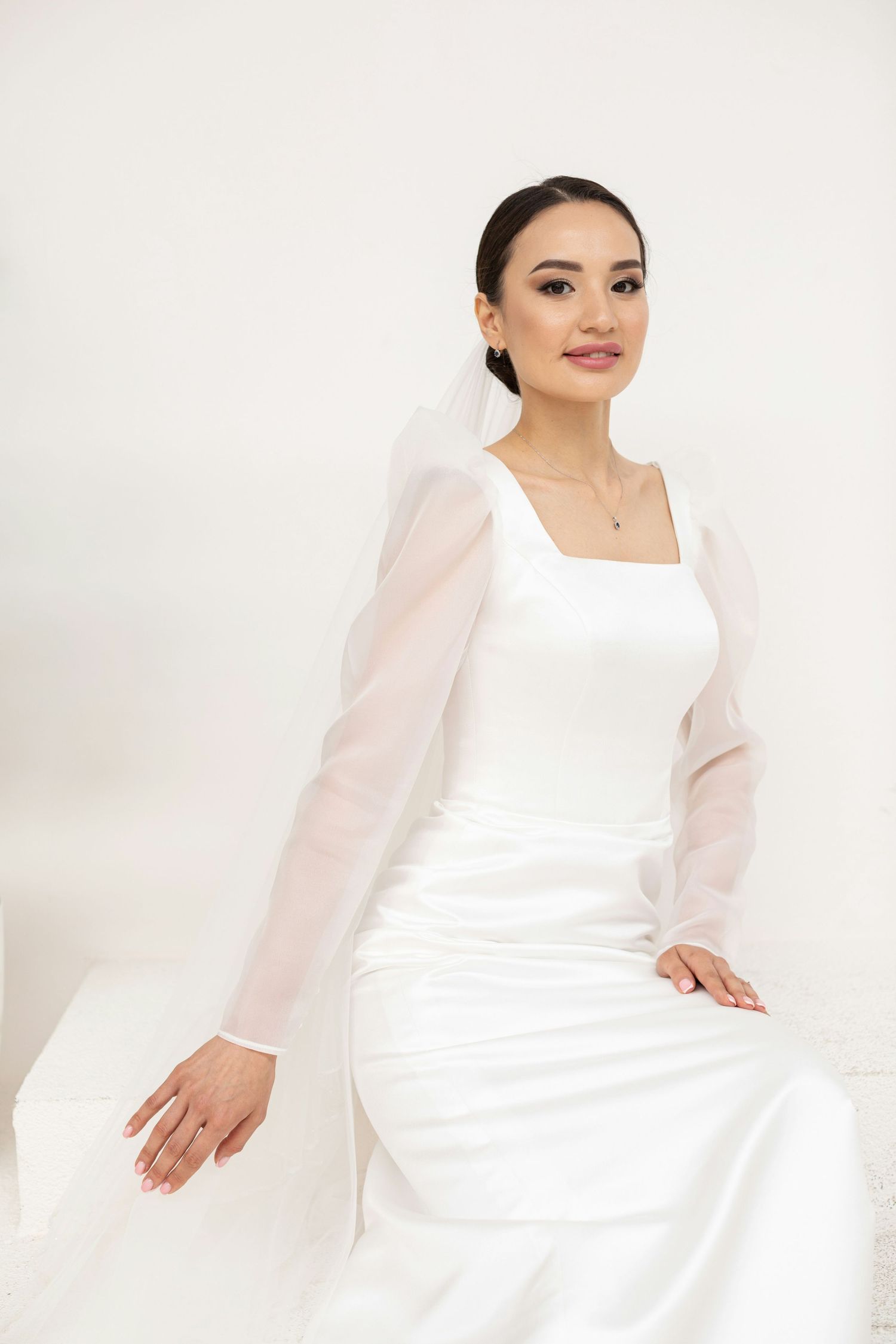 Sophisticated bride in satin square-neck gown with sheer organza sleeves seated on white surface.