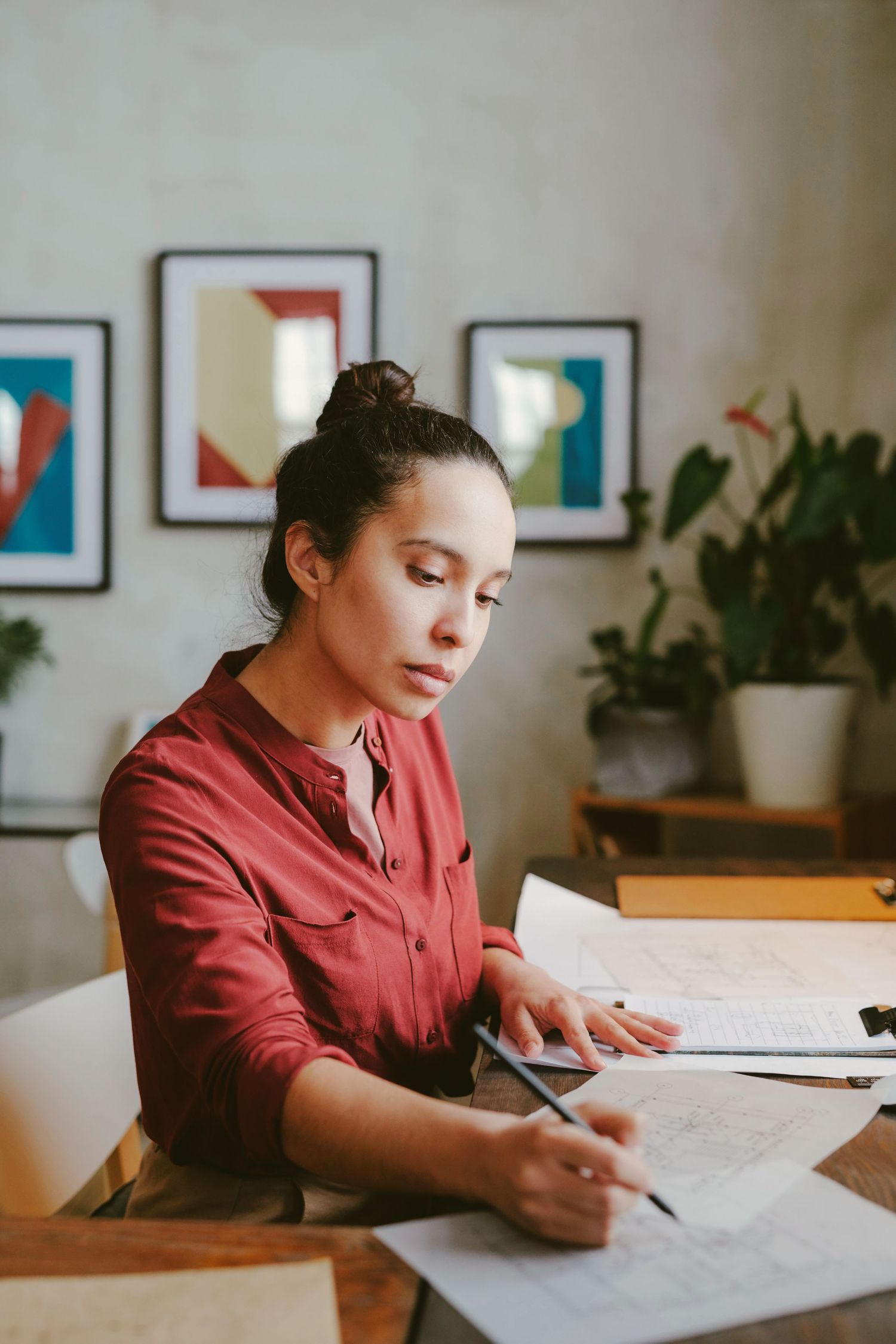 Woman in red shirt working at desk, reviewing architectural blueprints with framed art and plants in background.