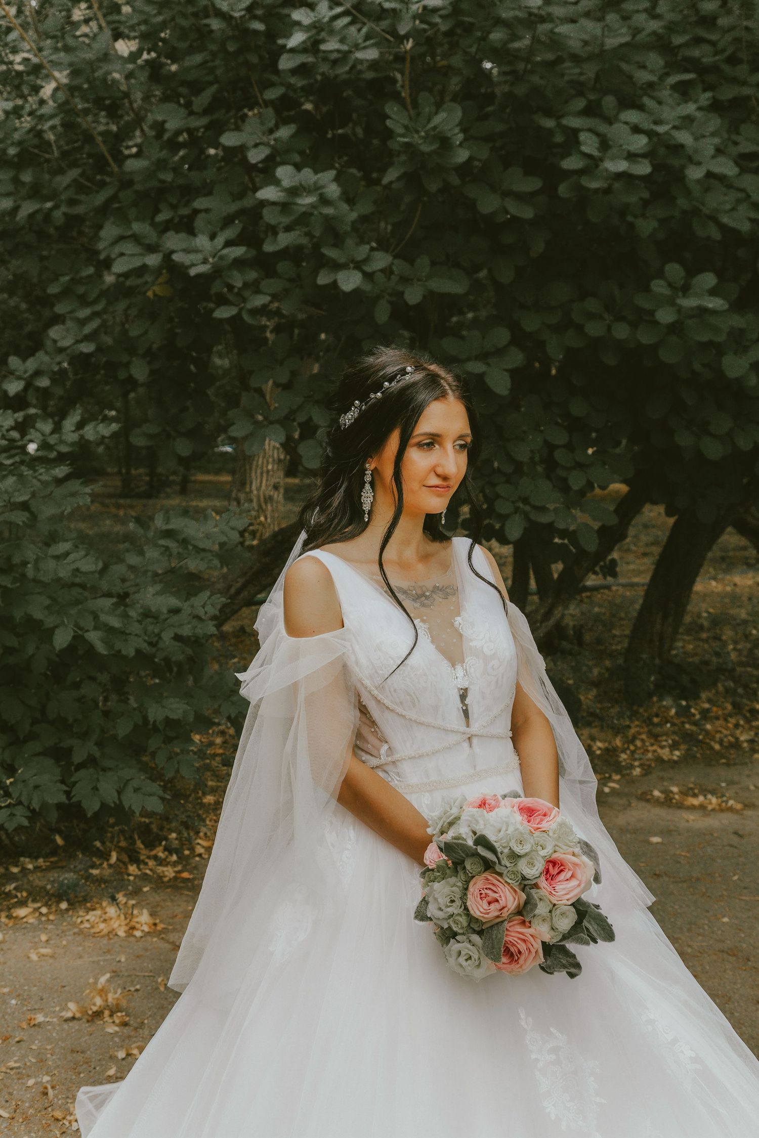 Bride in white off-shoulder gown holding pink bouquet, posing outdoors near lush green trees.