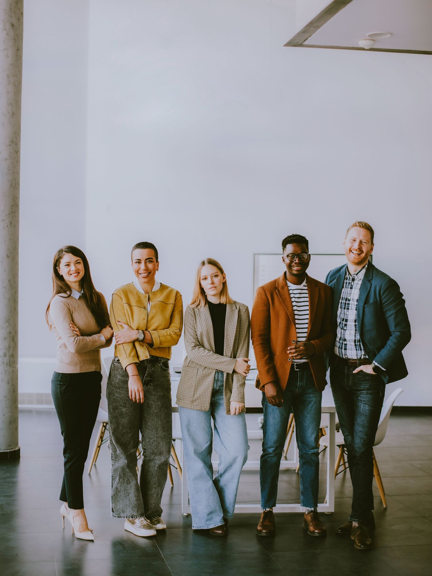 Five diverse young professionals smiling together in a bright modern office space, wearing casual stylish outfits.