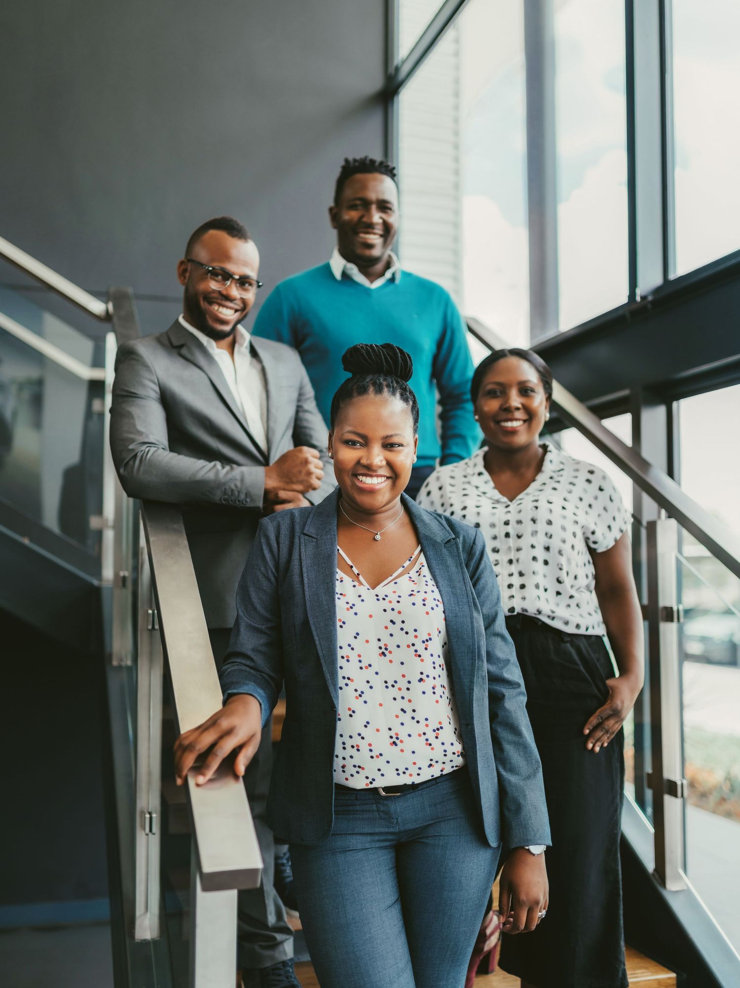 Four smiling Black business professionals posing together on modern office stairs in smart casual attire.