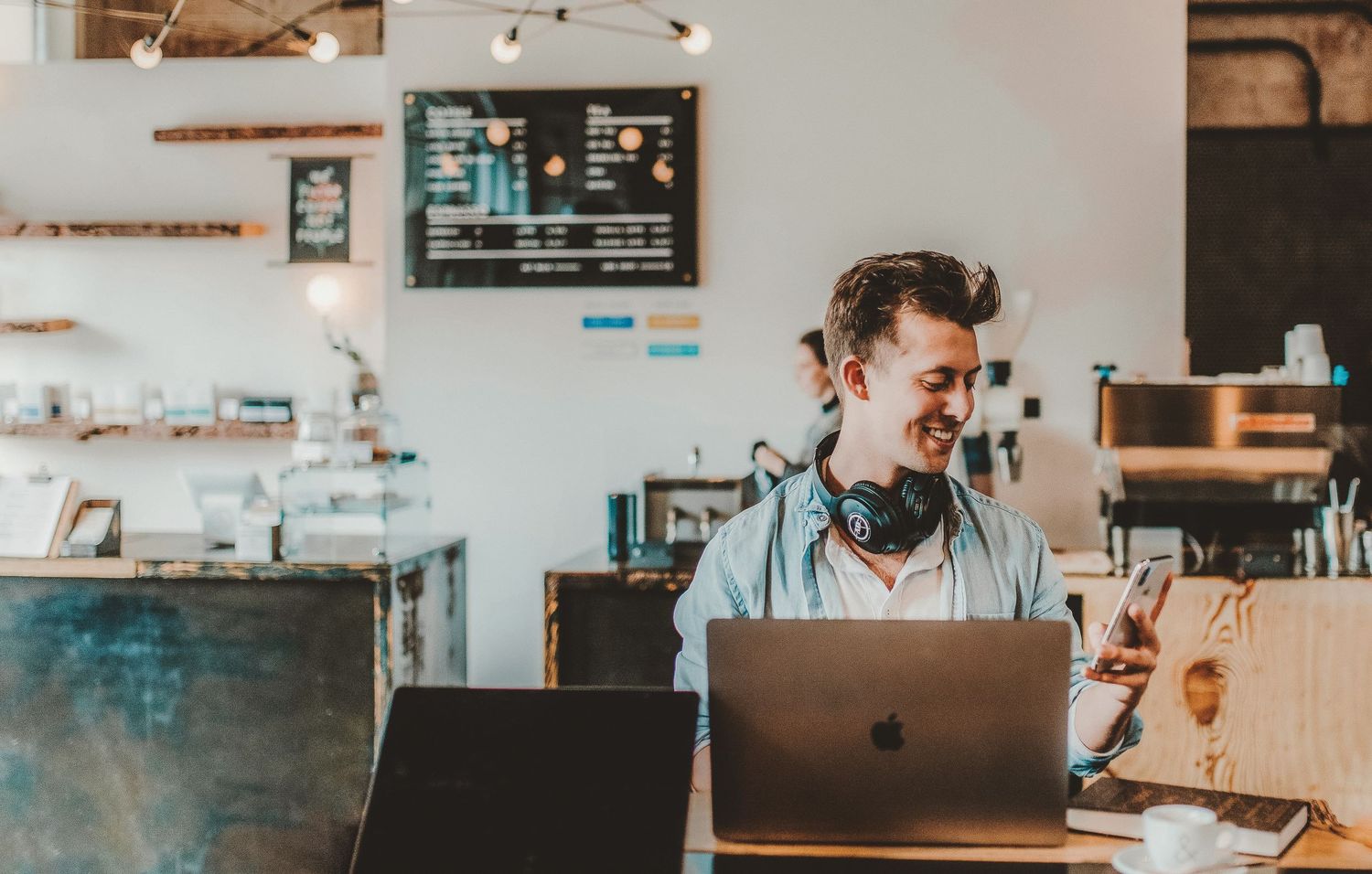 Smiling young man with headphones using MacBook laptop at counter in modern coffee shop.