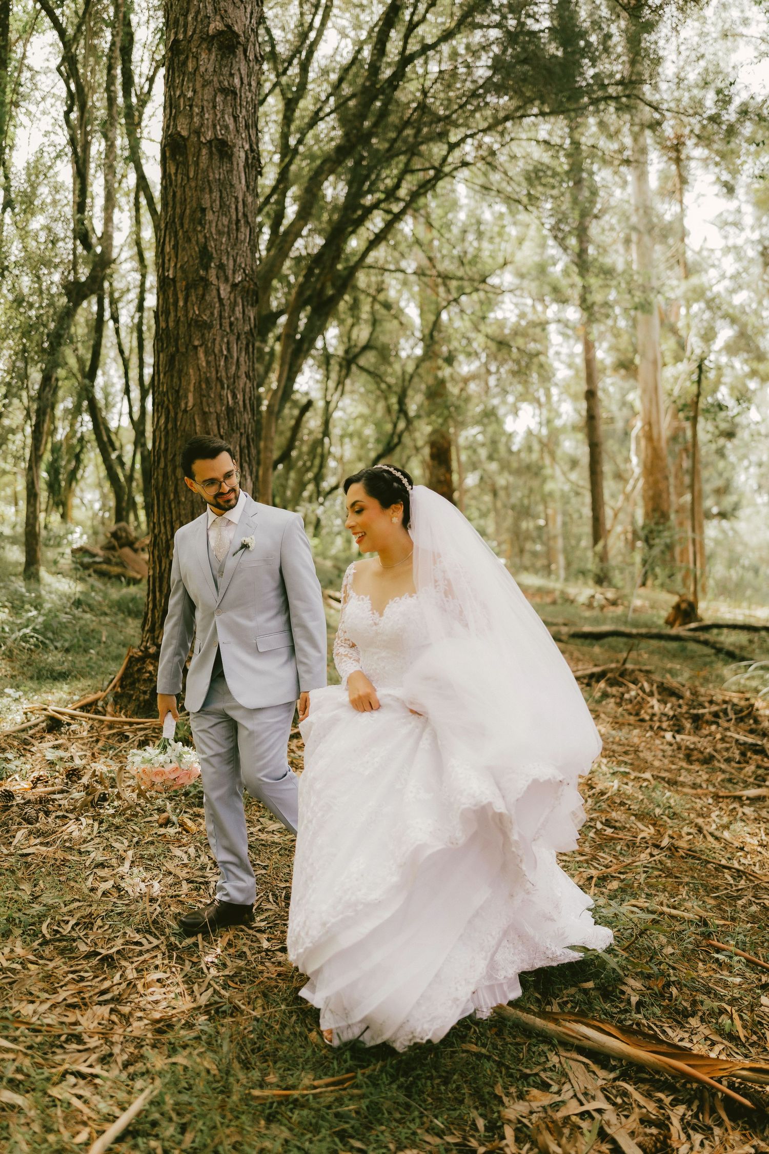 A bride in a white gown and veil walks hand-in-hand with a groom in a gray suit through a lush forest setting.