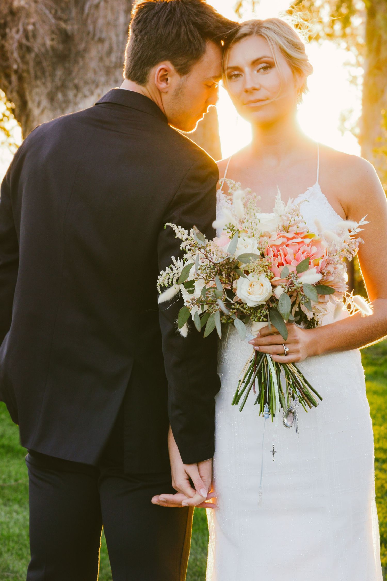 Bride holding pink and white bouquet embraces groom in black suit during golden hour outdoor wedding portrait.