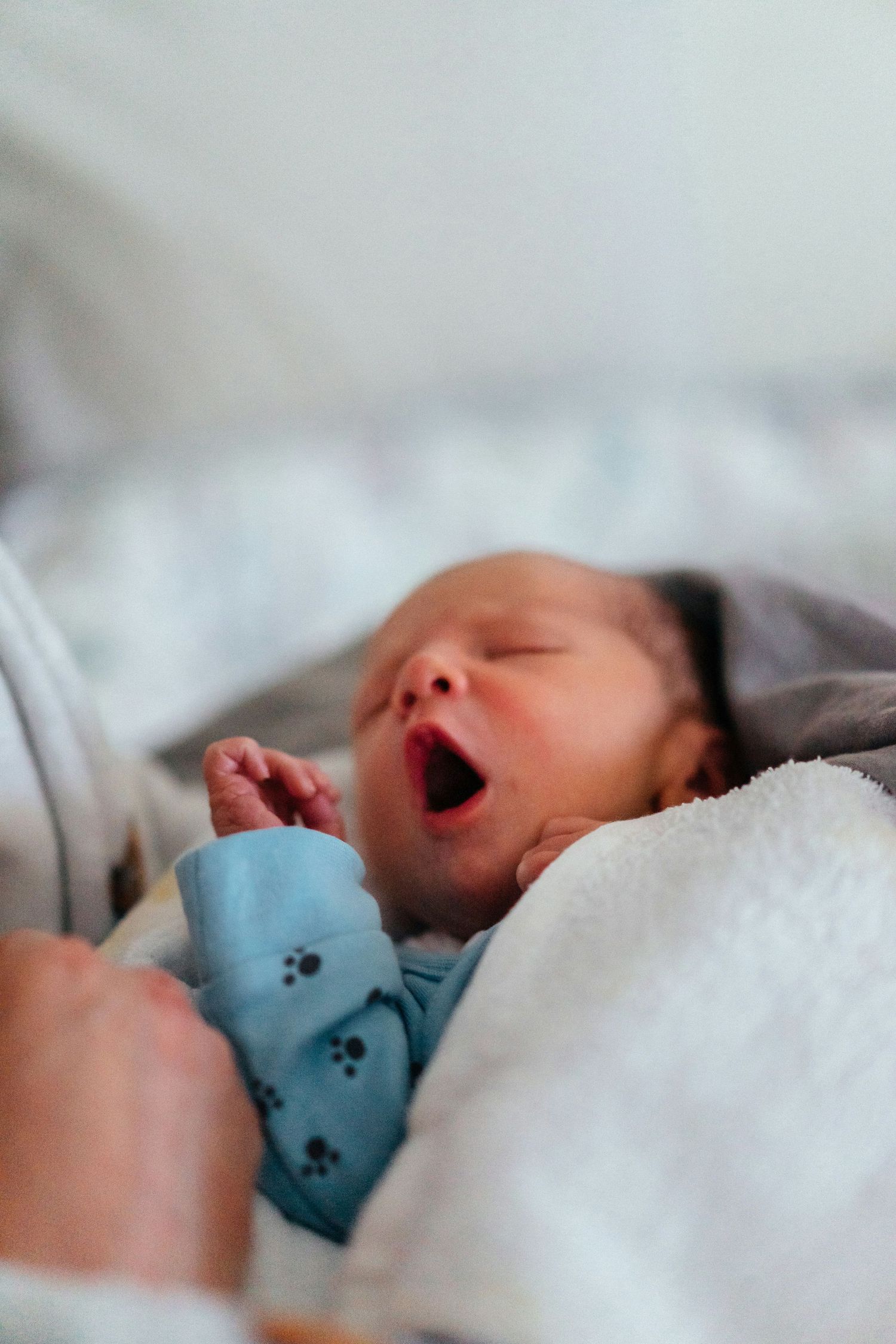 Newborn baby yawning while being held, wearing a blue onesie with paw prints in a hospital setting.