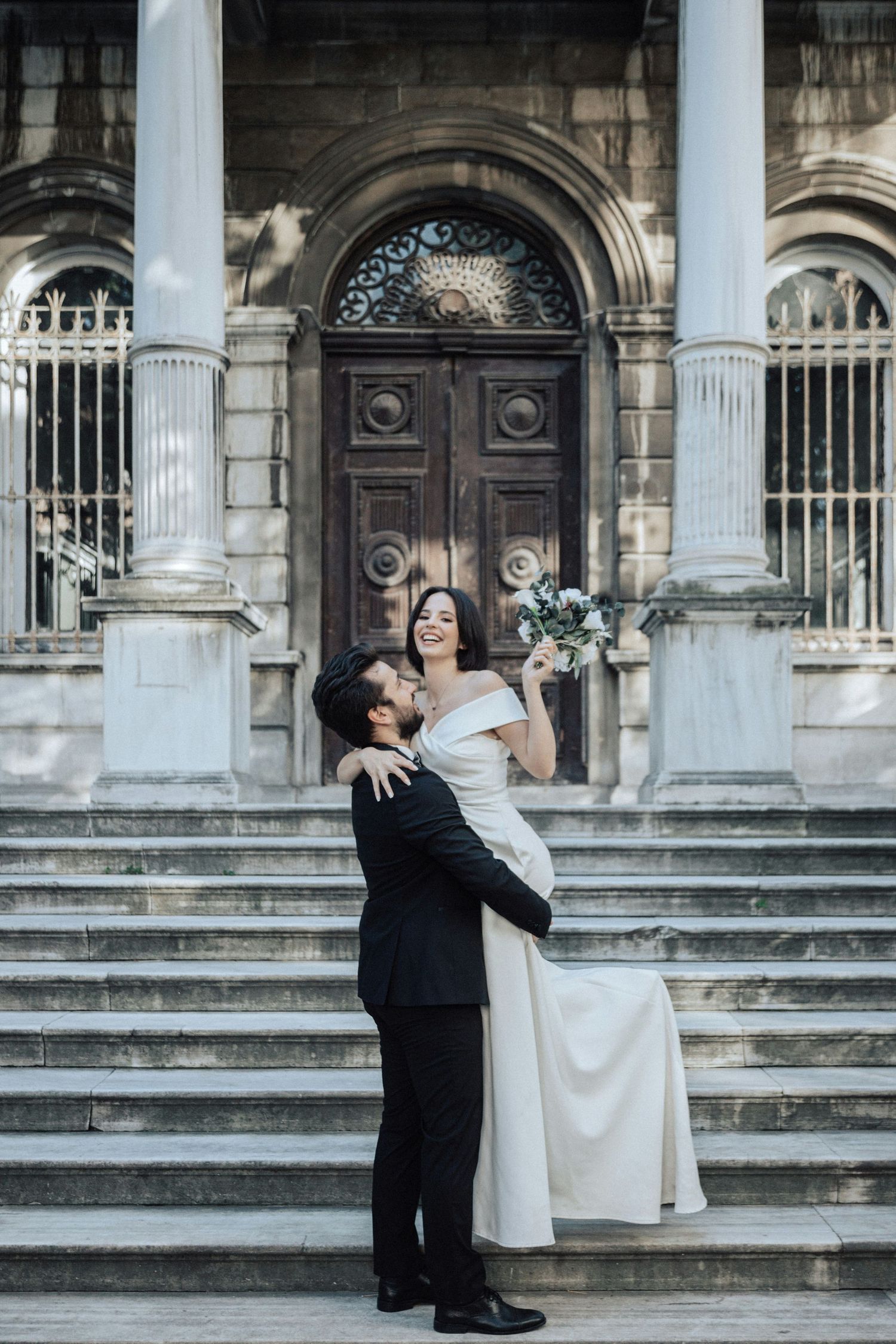 Groom lifts smiling bride in off-shoulder gown on steps of grand classical building with ornate wooden doors.